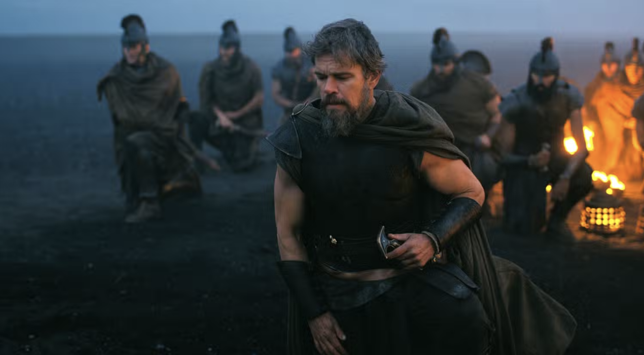 Bearded man in dark ancient warrior costume kneeling in front of a group of soldiers near a fire at dusk on a dark plain.