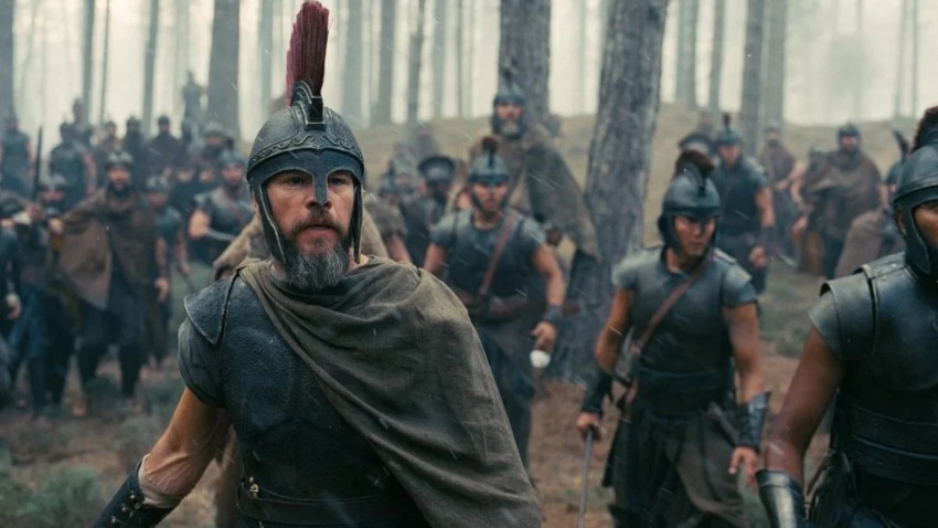 Bearded man in a red-plumed Greek helmet and cape running at the front of a large army of soldiers through a foggy forest.