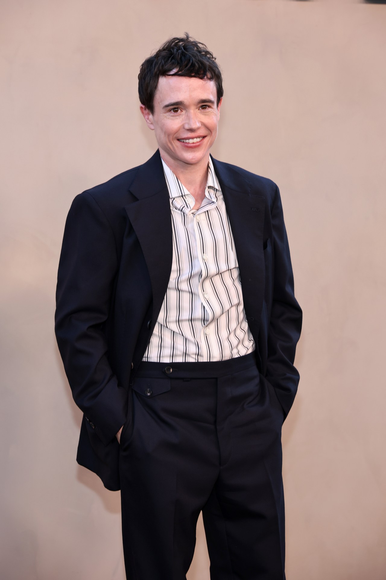 Elliot Page smiling in a dark suit and striped shirt at a movie premiere, standing against a beige backdrop.
