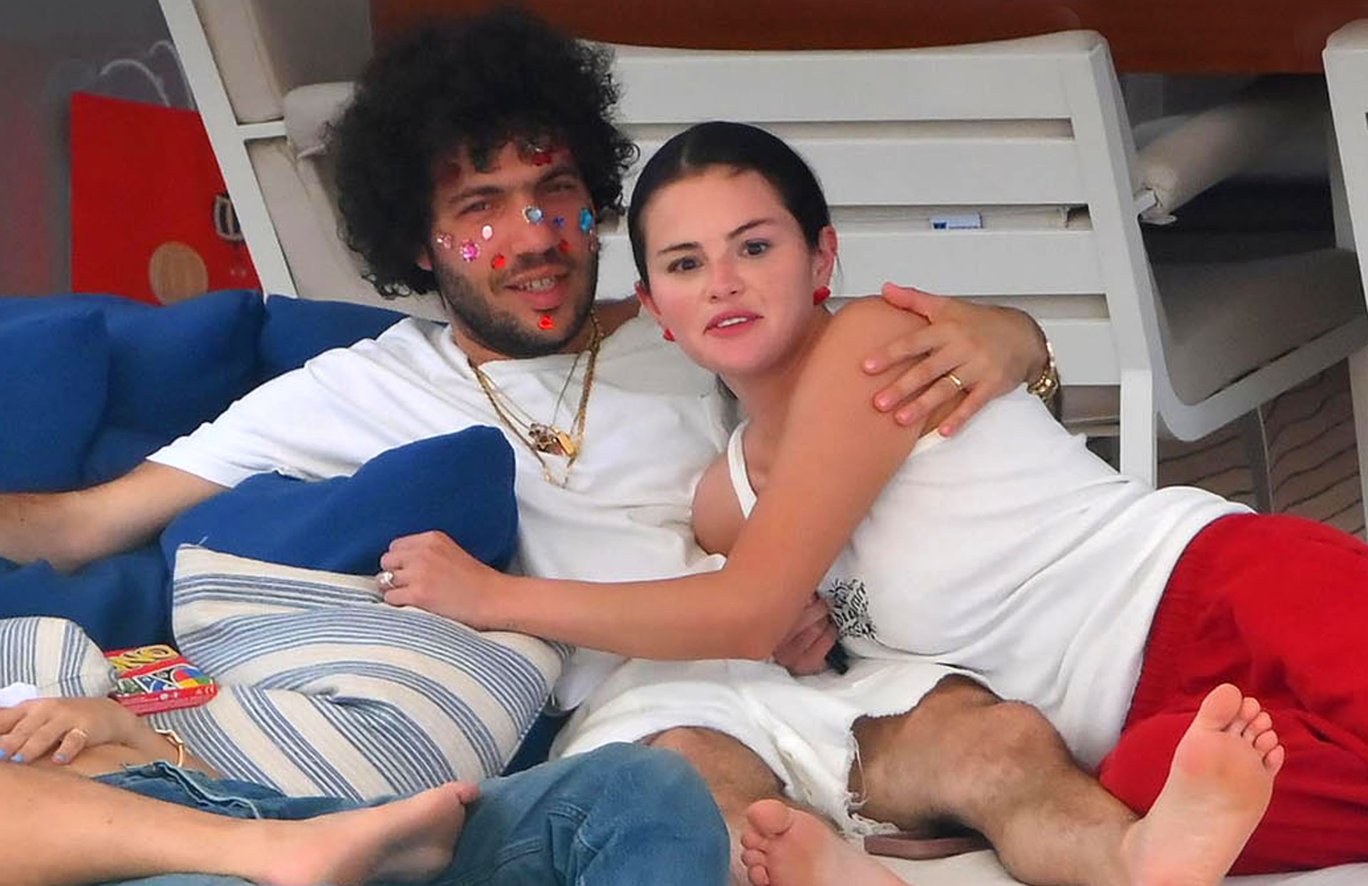Man with curly hair and colorful face stickers hugging a dark-haired woman in a white tank top on a boat lounge chair.