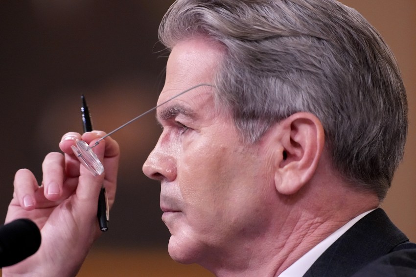 Close-up profile of a gray-haired man in a suit removing wire-rimmed glasses while holding a pen, facing left against a blurred brown background.