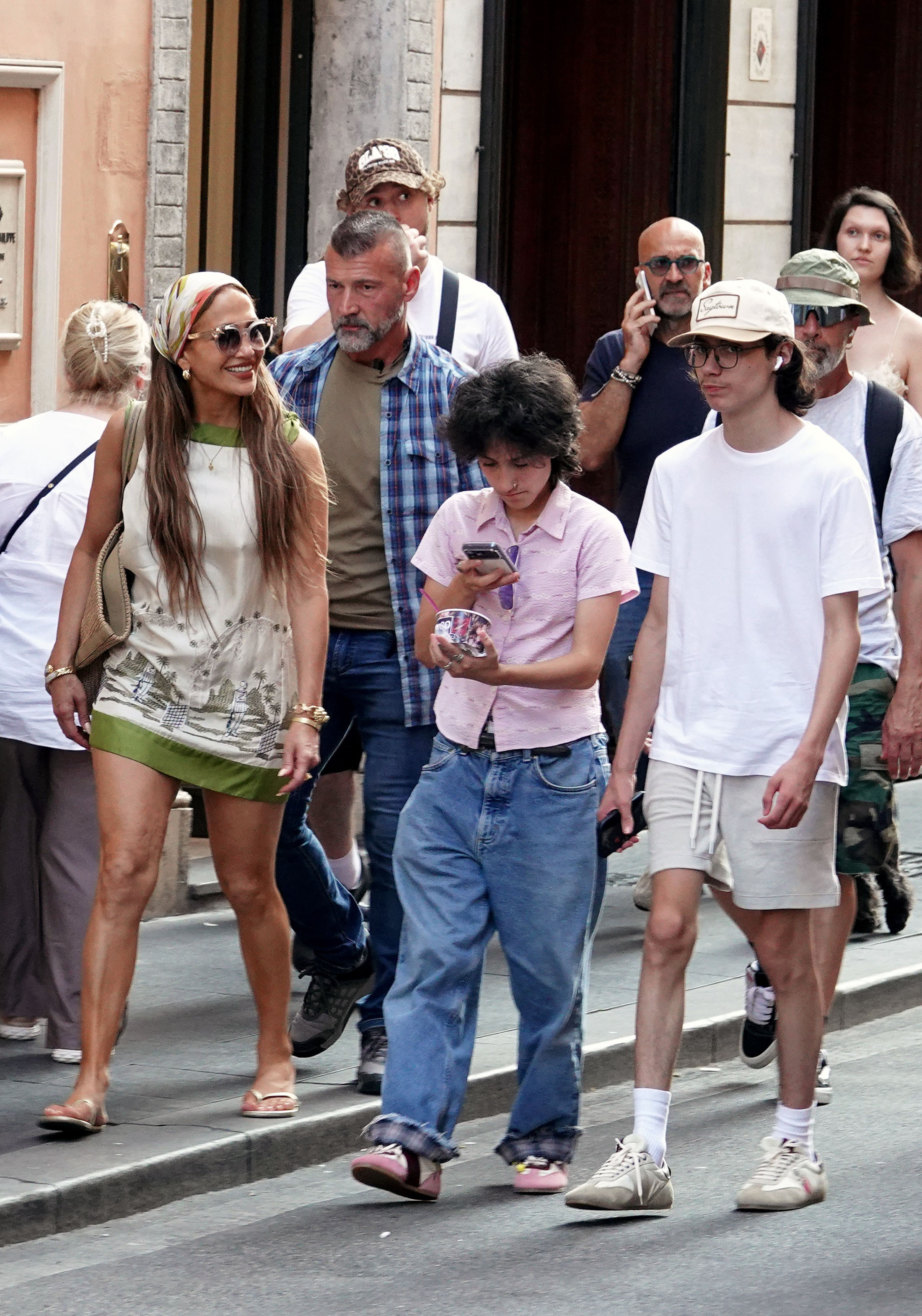 Jennifer Lopez smiles while walking on a busy Rome street with two teens — one in a pink shirt holding a phone and gelato, one in a white tee and shorts — surrounded by a crowd.