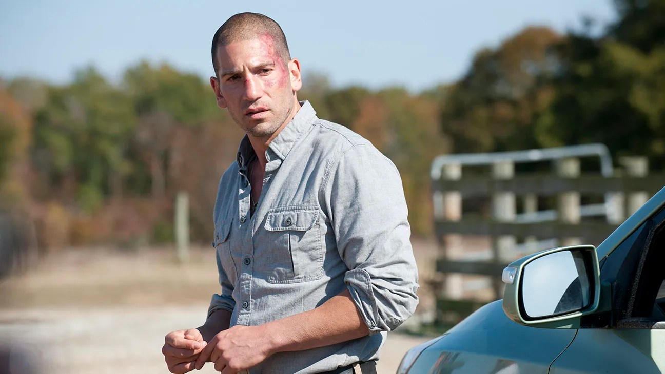 Short-haired man with cuts and bruises on his face leaning against a car door outdoors, looking back with a tense expression.