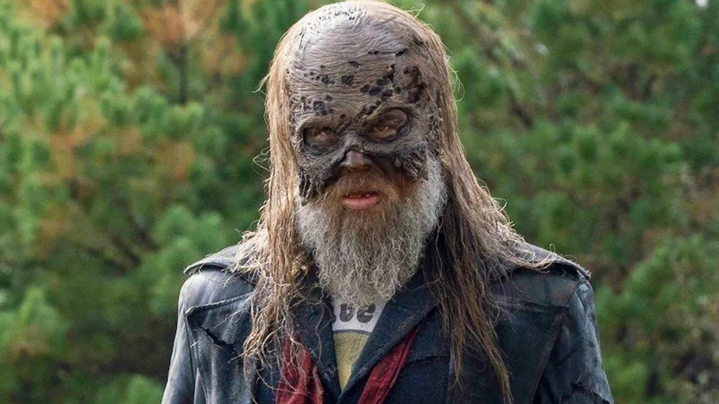 Person wearing a decayed zombie-face mask with long grey hair and beard, standing in front of green pine trees.