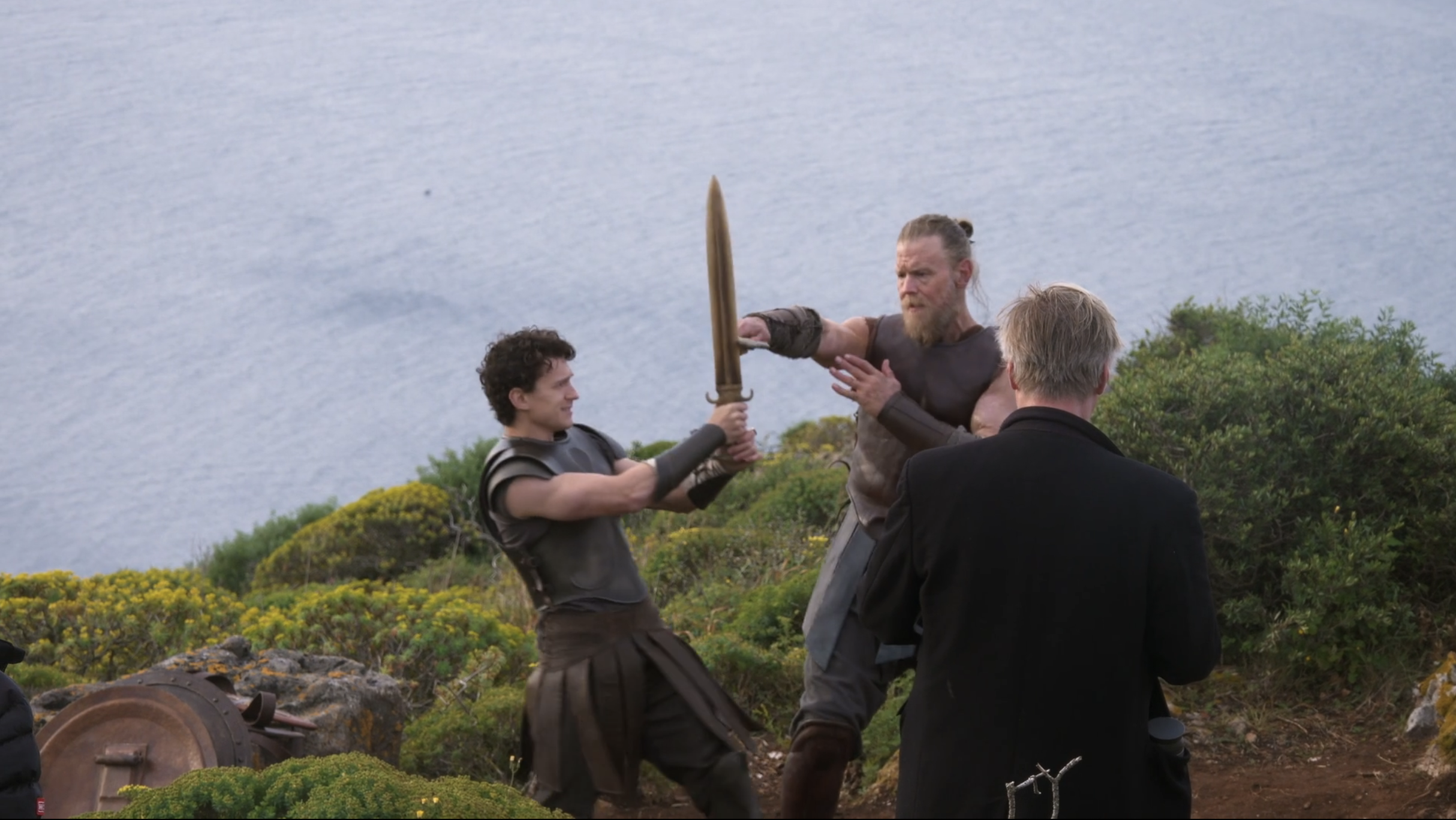 Two men in ancient warrior costumes clashing with a bronze sword on a rocky coastal hillside, with a crew member nearby.