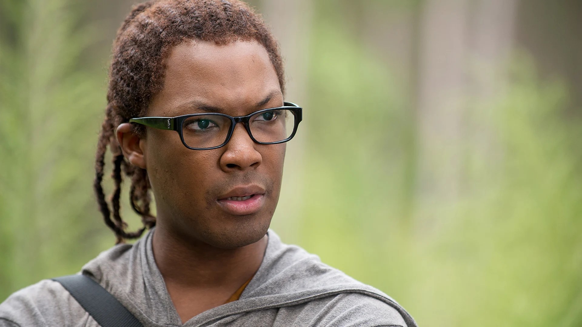 Close-up of a young man with locs and black rectangular glasses wearing a gray hoodie, standing outdoors in a wooded setting.