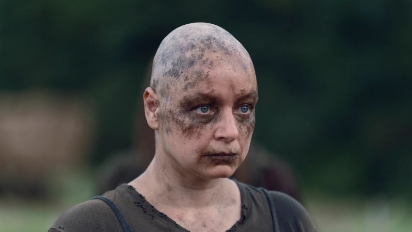 Close-up of a shaved-head woman with dirt and grime makeup covering her face, wearing a torn dark shirt, against a blurred outdoor background.