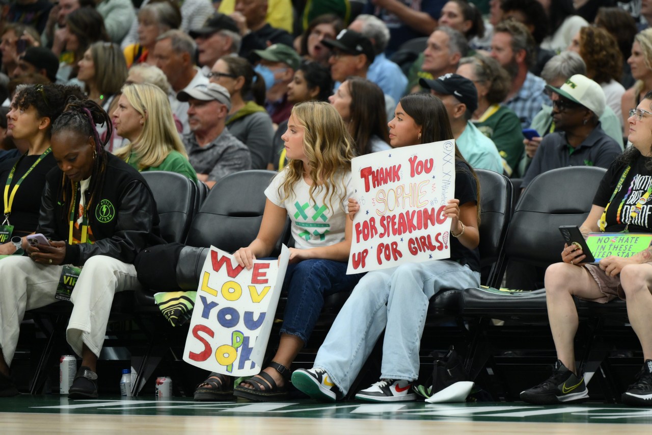 Two teenage girls sitting courtside hold handmade signs reading 'We Love You Soph' and 'Thank You Sophie For Speaking Up For Girls' at a WNBA game.