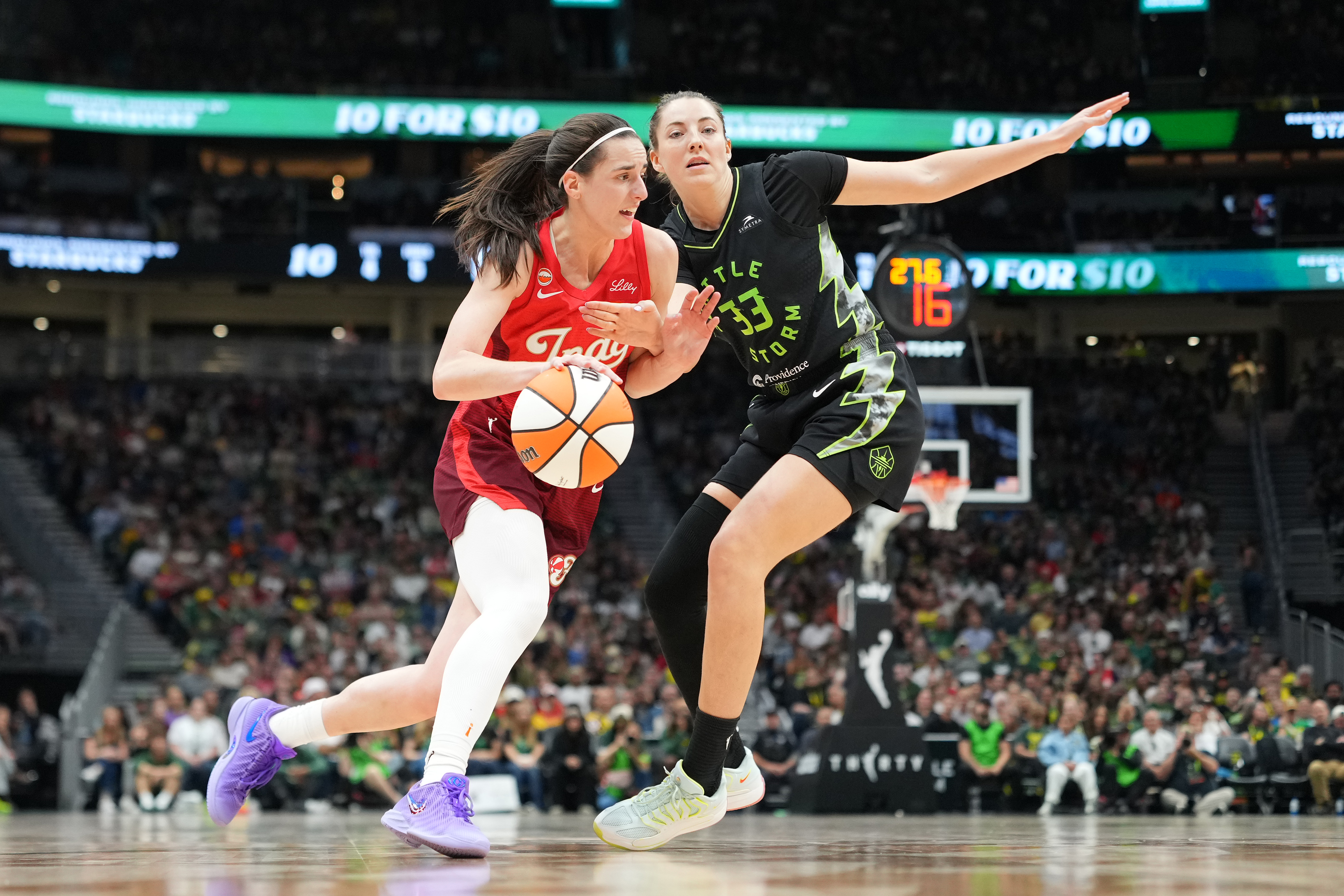 Caitlin Clark in a red Indiana Fever jersey dribbles past Seattle Storm defender Katie Lou Samuelson (#33) on the court at Climate Pledge Arena.