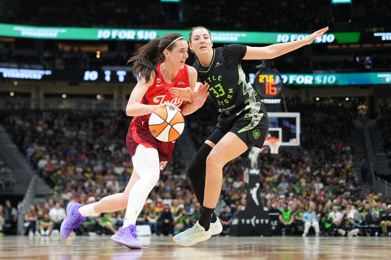 Caitlin Clark in a red Indiana Fever jersey dribbles past Seattle Storm defender Katie Lou Samuelson (#33) on the court at Climate Pledge Arena.