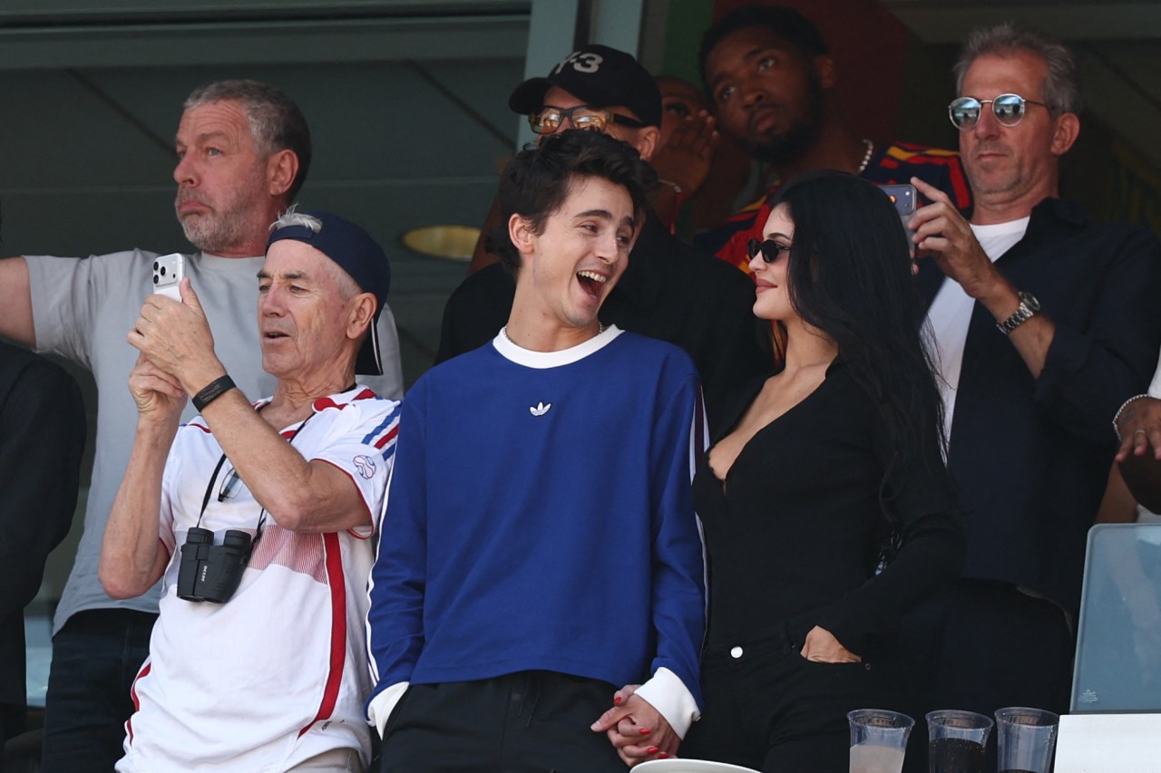Timothée Chalamet laughing and holding hands with Kylie Jenner in stadium suite seating, with an older man taking a photo beside them.