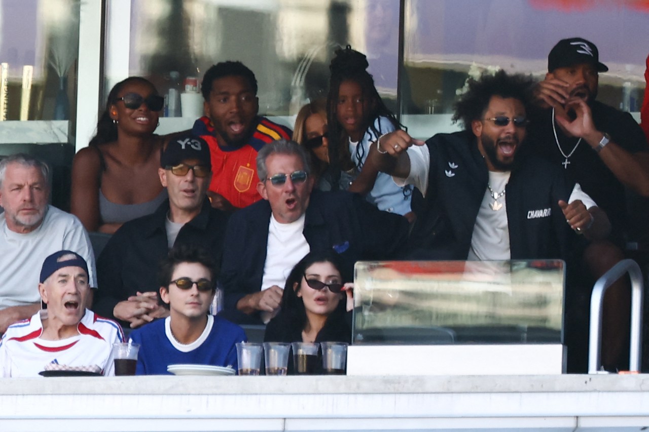 A crowded stadium suite with multiple people reacting emotionally to a goal, including a young man in a blue Adidas sweatshirt and a dark-haired woman in sunglasses in the front row.