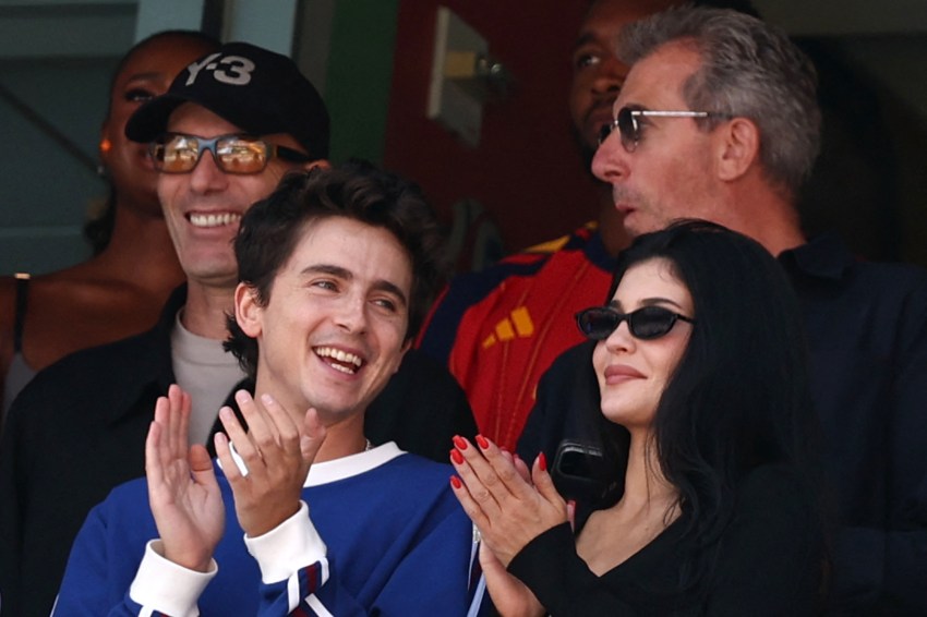 Timothée Chalamet clapping and laughing next to Kylie Jenner wearing black sunglasses at a stadium suite