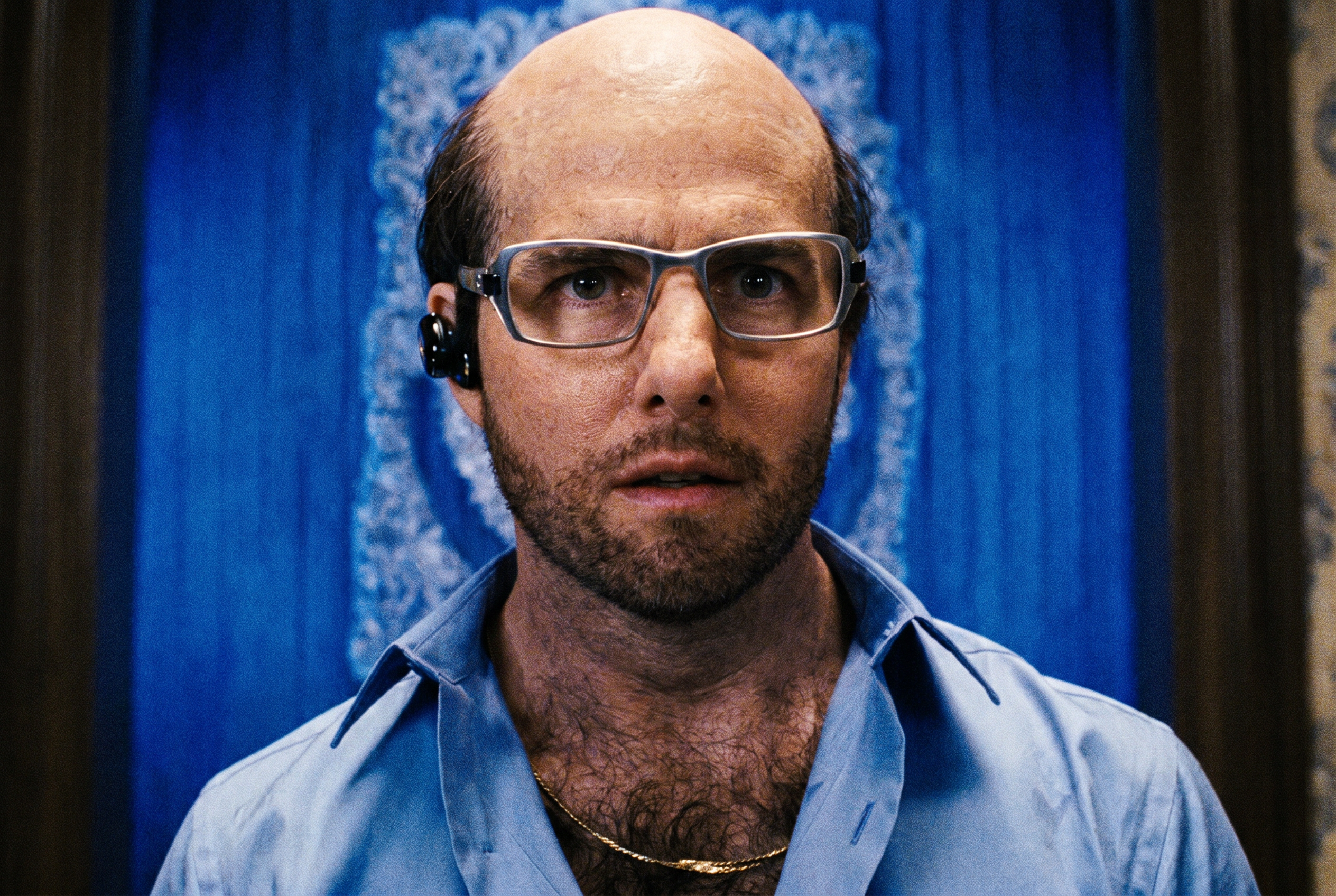A bald man with glasses, a stubbled beard, a gold chain, and a Bluetooth earpiece wearing an open blue shirt, heavy prosthetic makeup.