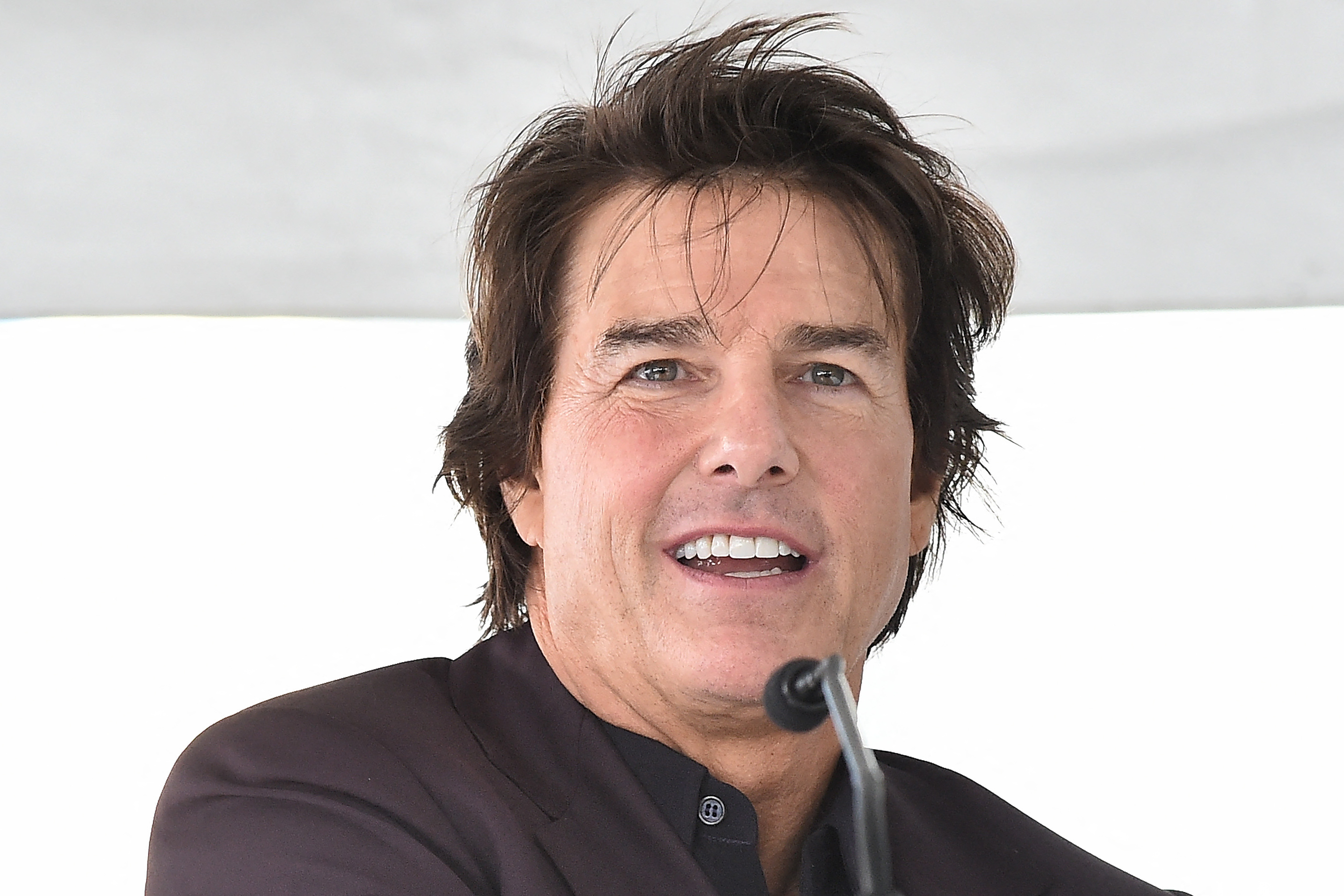 Tom Cruise smiling and speaking into a microphone at an outdoor event, wearing a dark jacket.