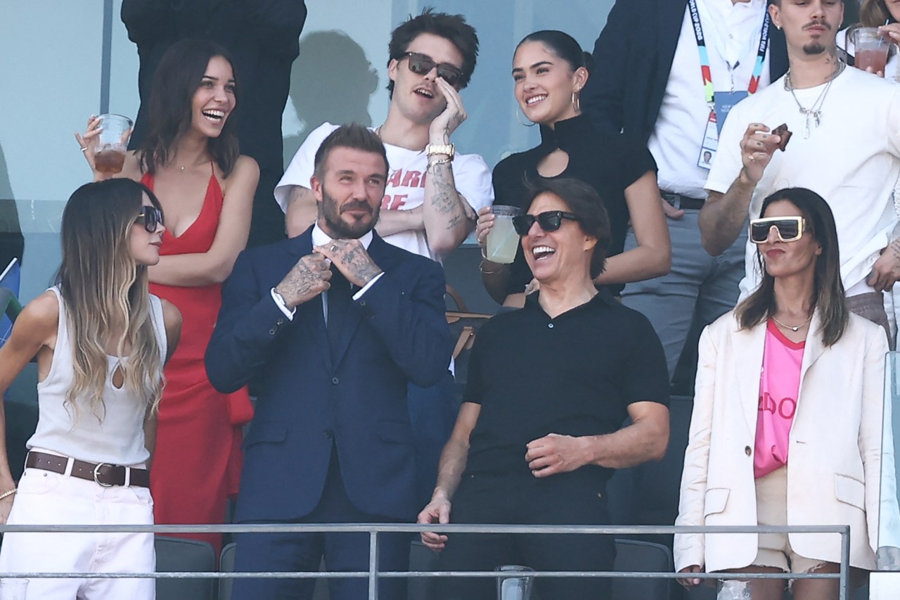 David Beckham in a navy suit, Tom Cruise in black, and Victoria Beckham in white laugh together in a crowded VIP stadium box.