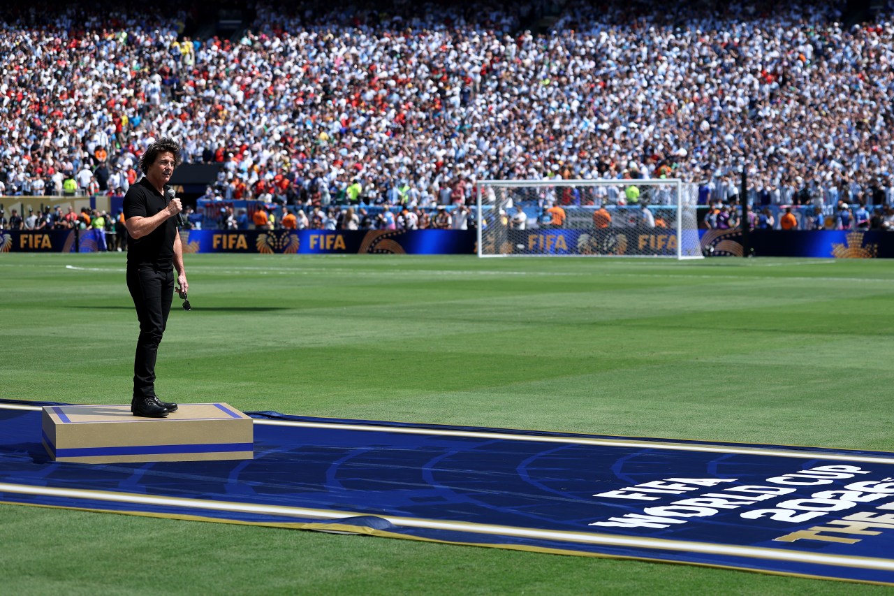 A man in all black stands on a raised platform on a soccer field holding a microphone, with a massive stadium crowd and FIFA branding visible behind him.