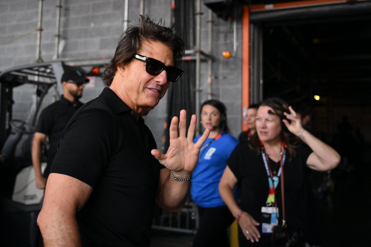 A man in a black polo shirt and dark sunglasses waves with one hand while walking through a backstage stadium area, with event staff visible behind him.