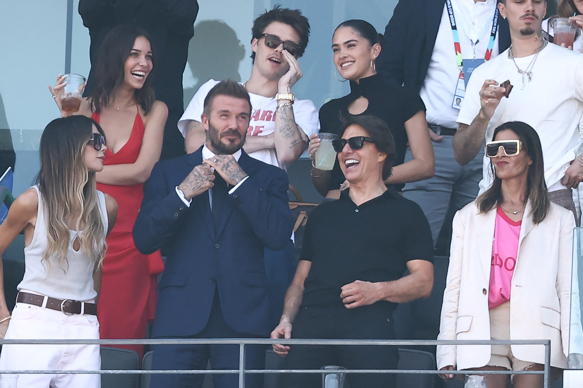 David Beckham in a navy suit, Tom Cruise in black, and Victoria Beckham in white stand laughing in a stadium VIP box, with several younger adults behind them.