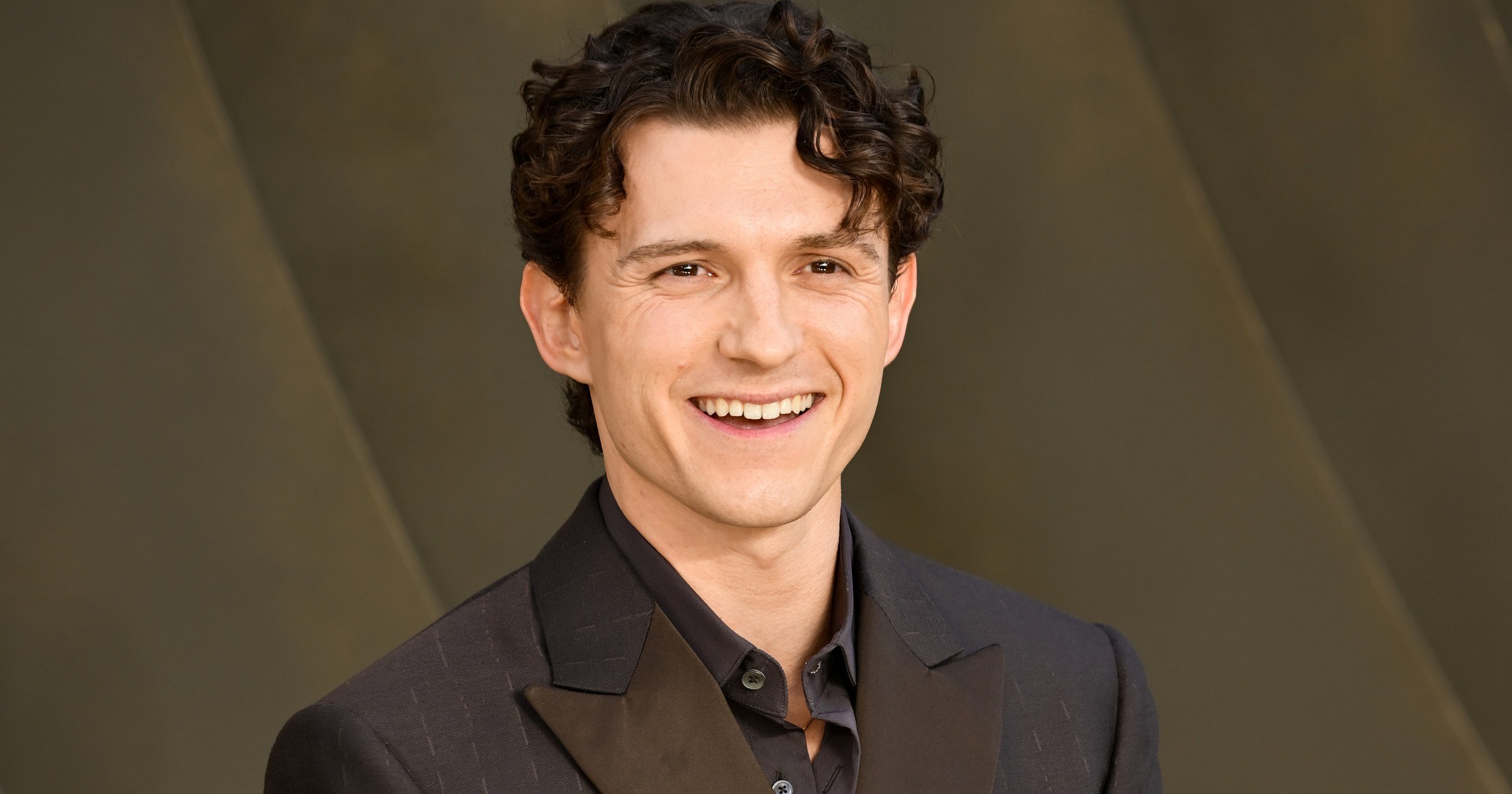 Tom Holland smiling in a dark double-breasted pinstripe suit with hands clasped, posed against a dark gold abstract backdrop.