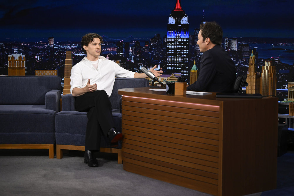 A young man in a white shirt sits with one arm outstretched toward the host's desk on The Tonight Show set, animatedly telling a story.