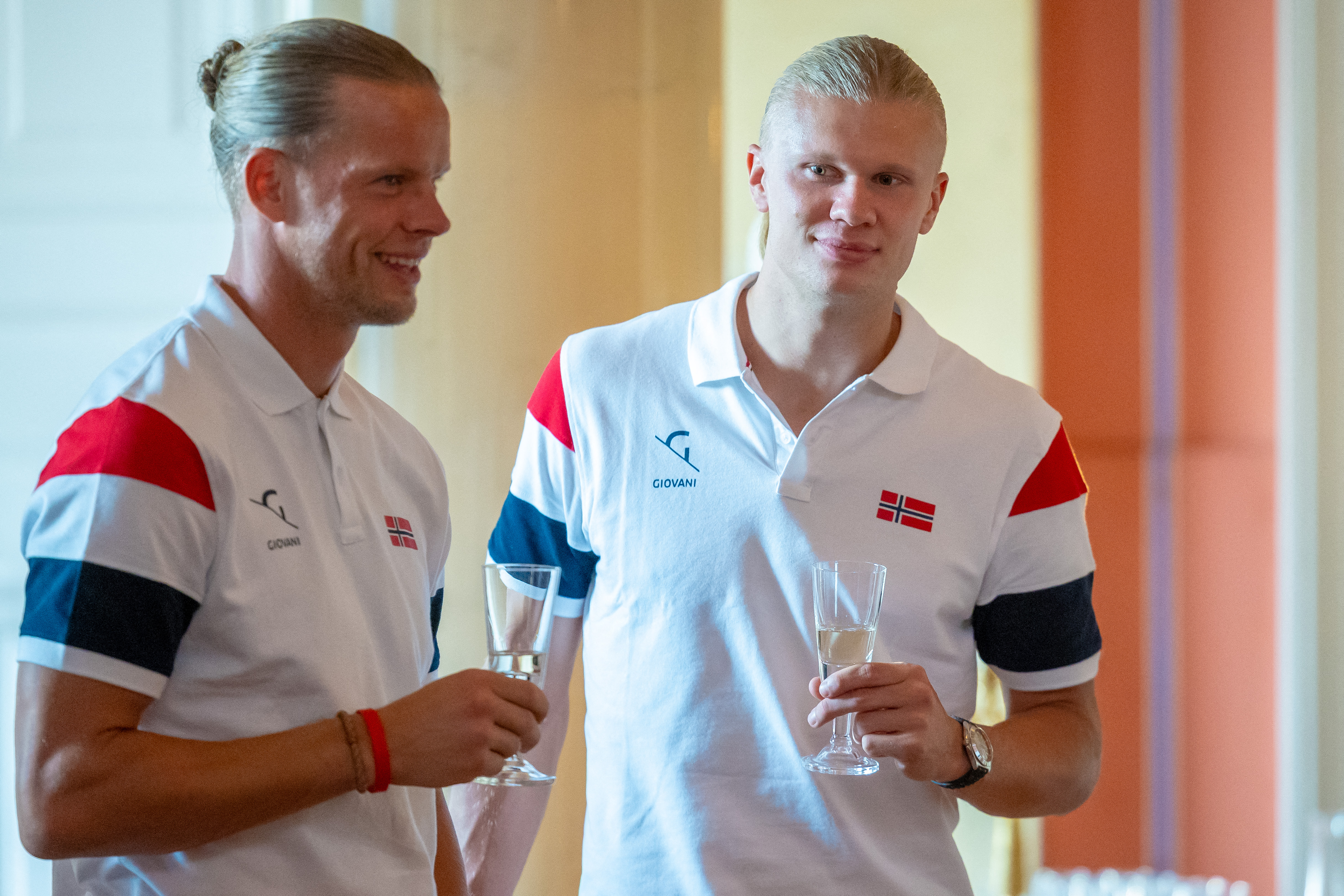 Two tall blond men in matching white Norway Giovani polo shirts hold champagne flutes and chat at an indoor reception.