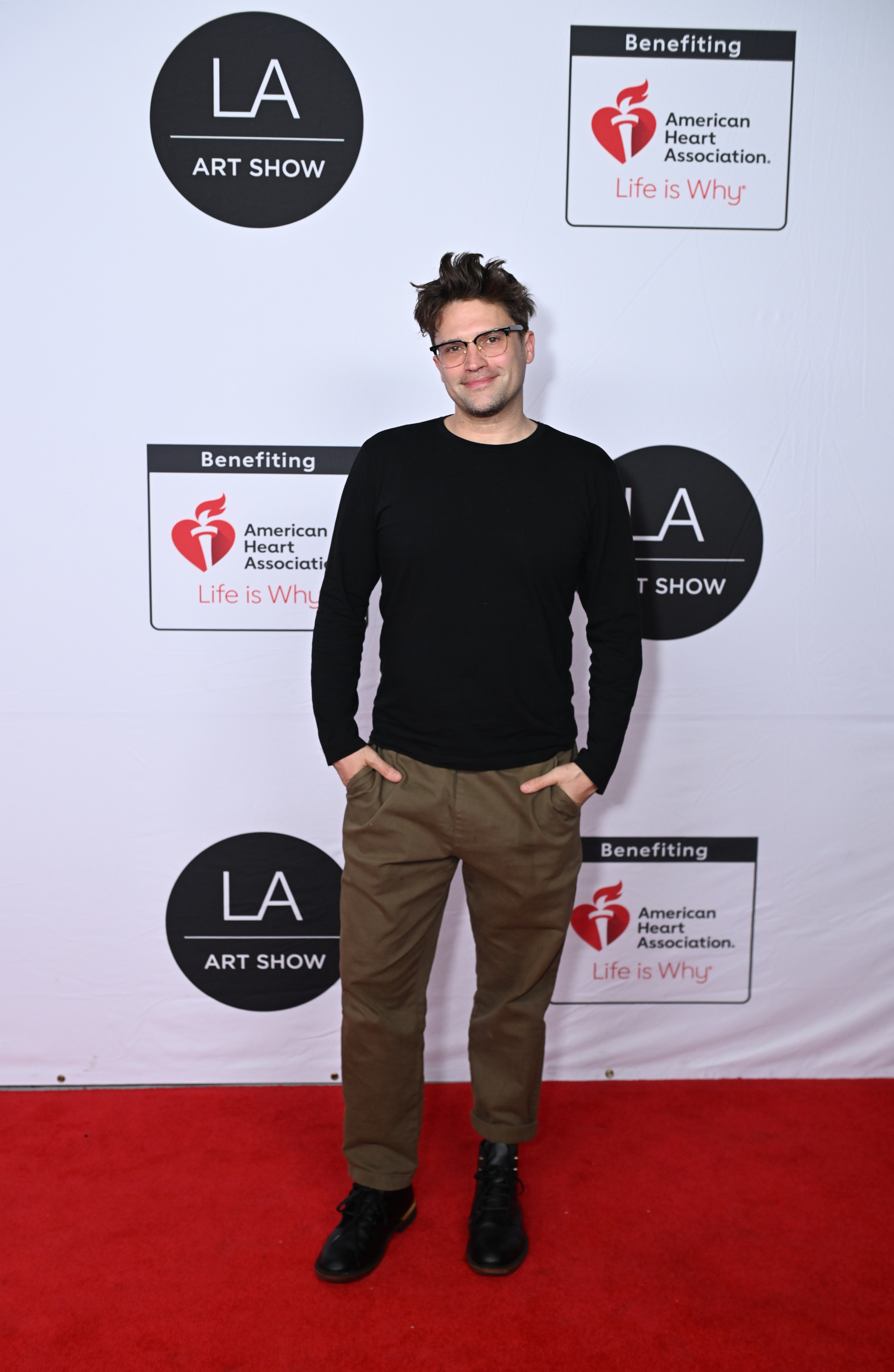 Tom Schwartz in a black long-sleeve shirt, olive pants, and black boots standing on a red carpet in front of an LA Art Show step-and-repeat banner.