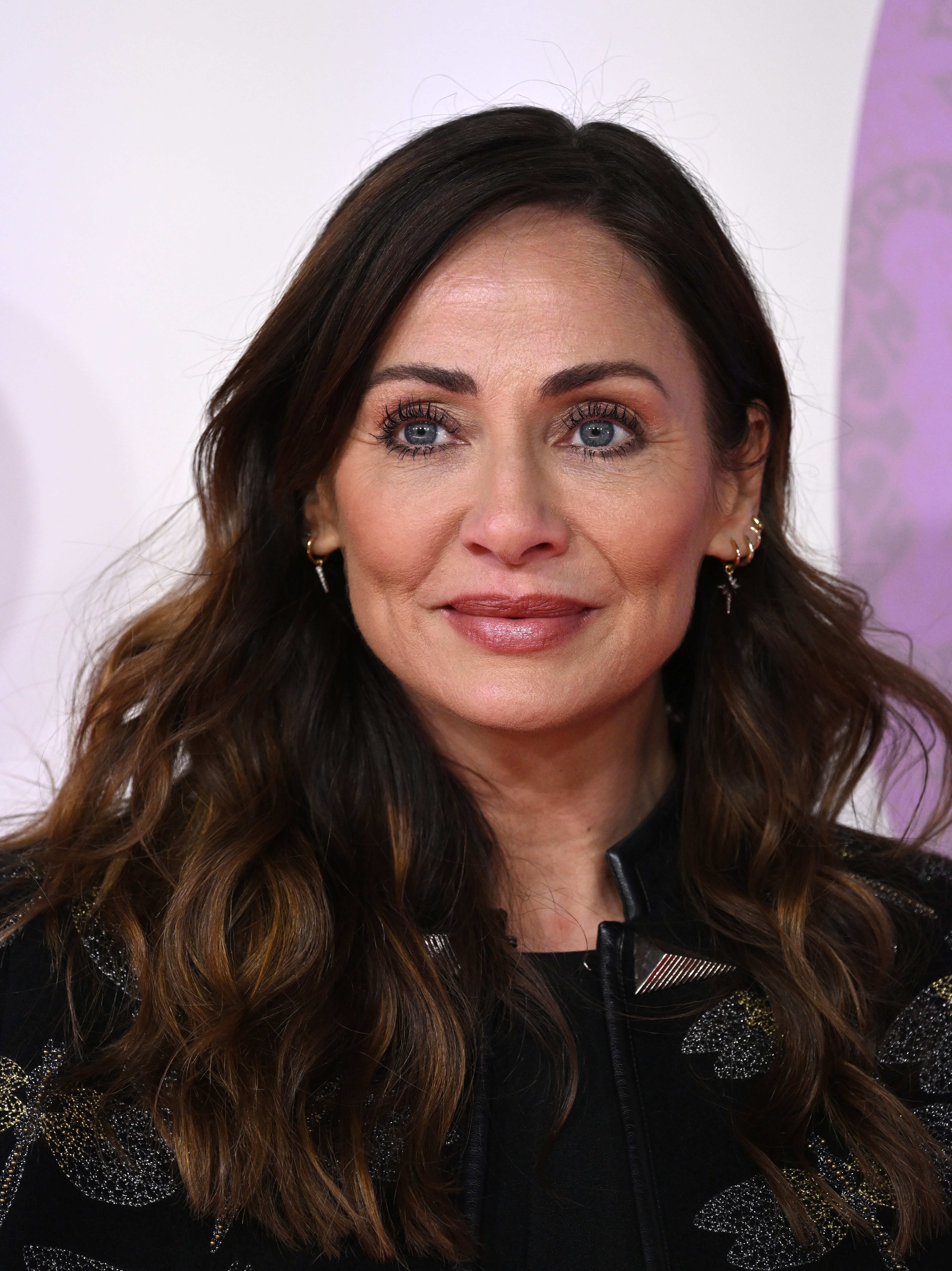 Woman with long dark wavy hair and blue eyes smiling at a film premiere, wearing a black embroidered jacket and gold hoop earrings.