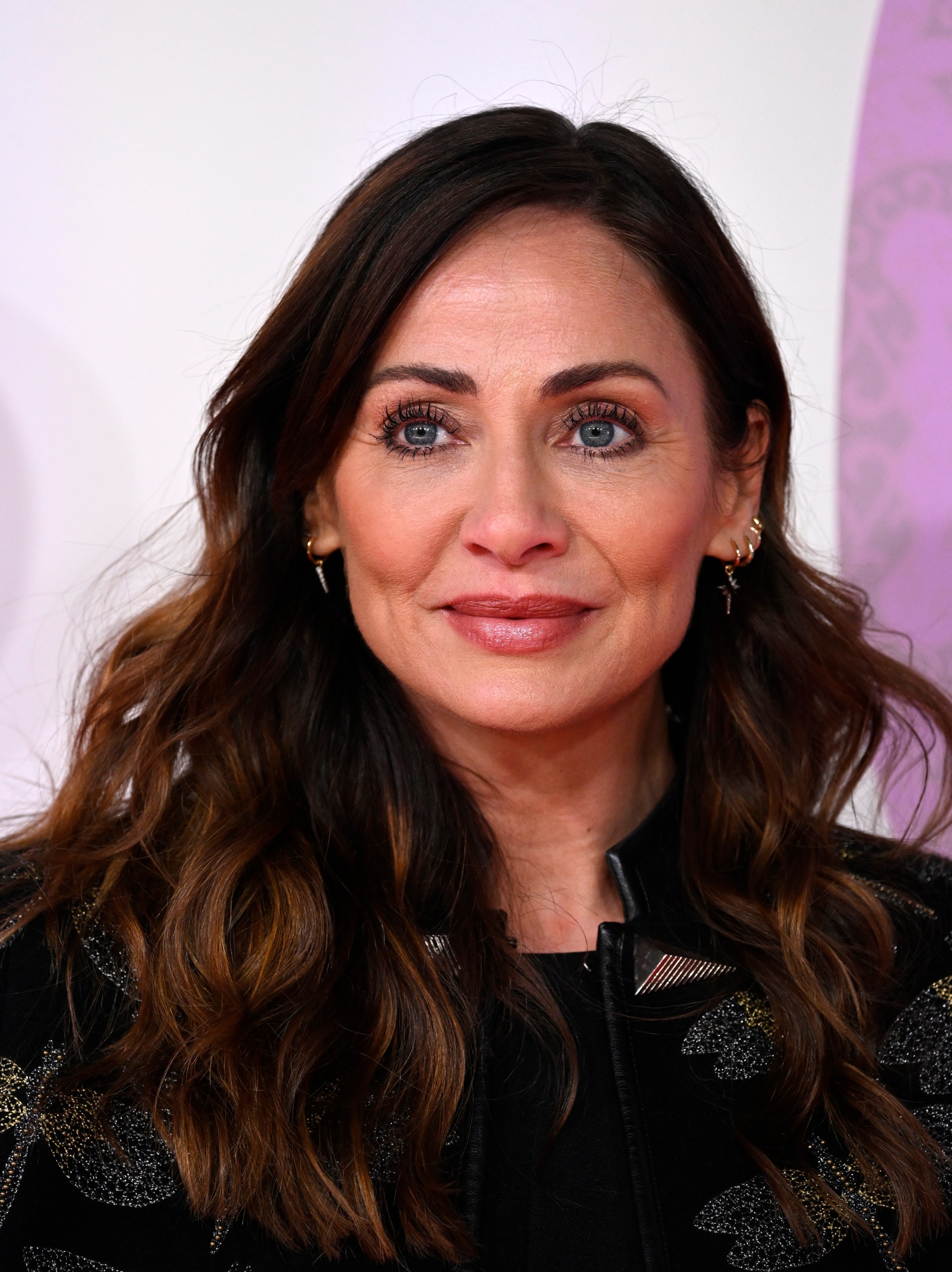 Woman with long dark wavy hair and blue eyes smiling at a film premiere, wearing a black embroidered jacket and gold hoop earrings.