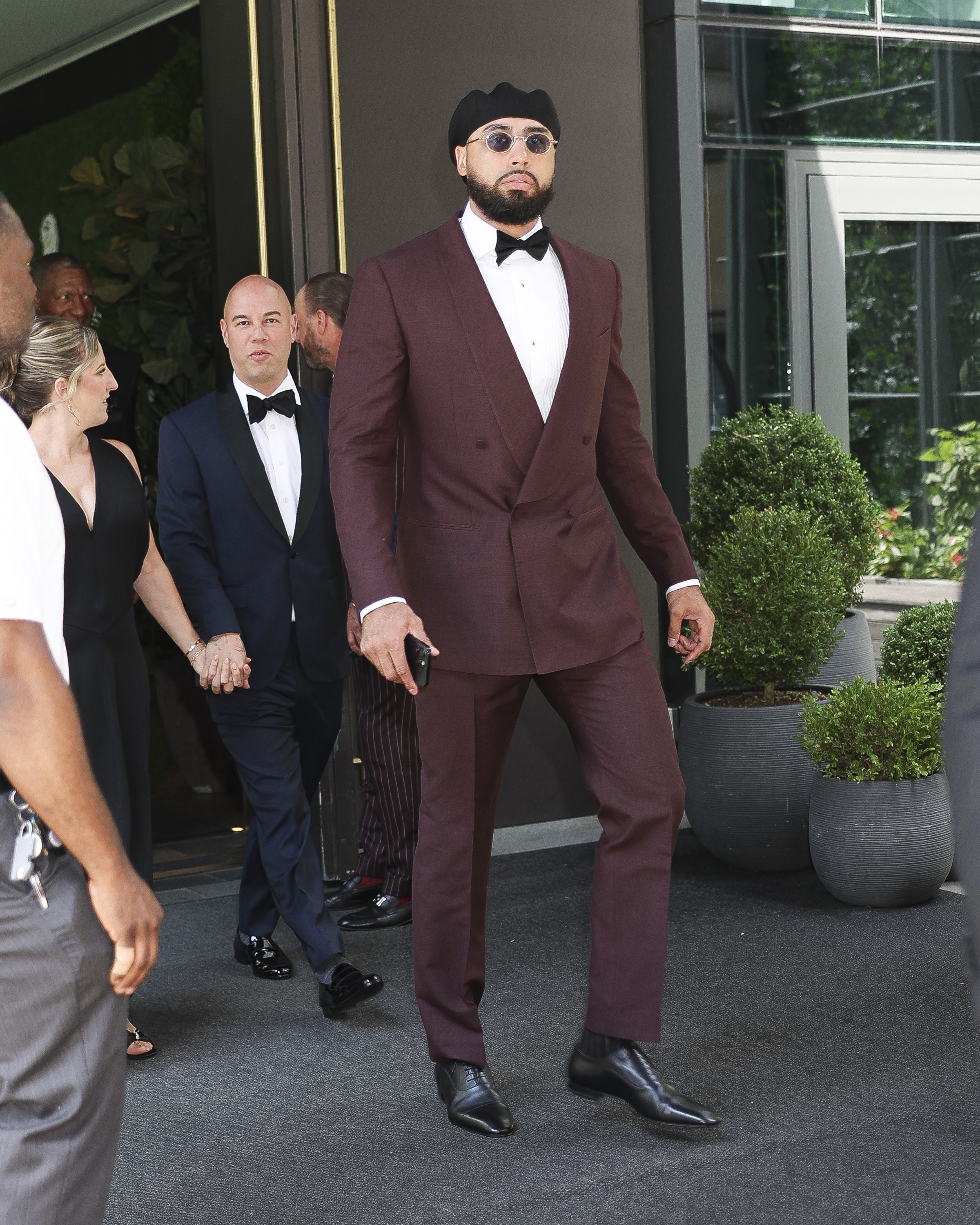 Tall man in a burgundy double-breasted suit, black bow tie, black cap, and round sunglasses walking outside a building in New York City.
