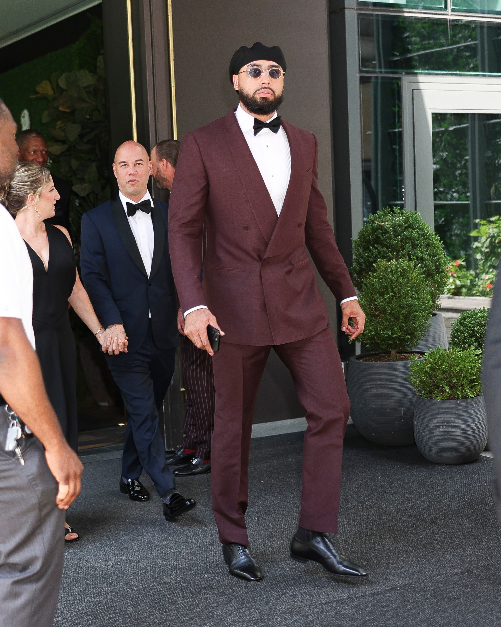 Tall man in a burgundy double-breasted suit, black bow tie, black cap, and round sunglasses walking outside a building in New York City.