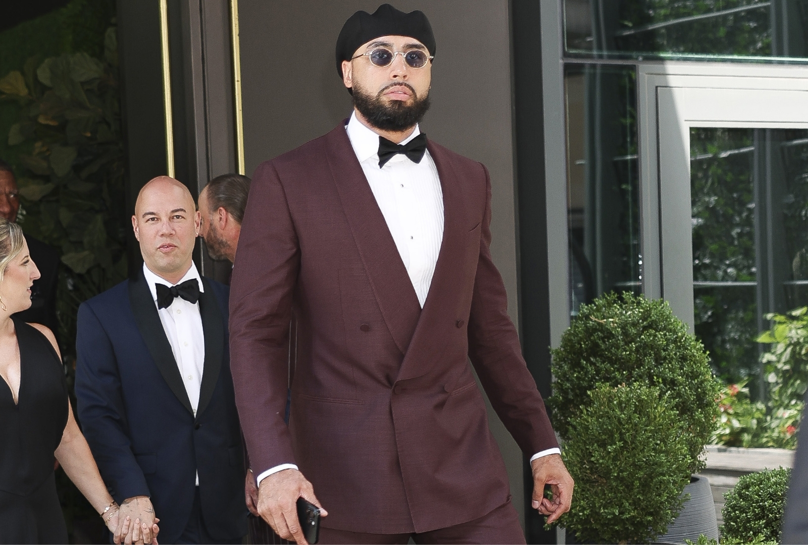 Tall man in a burgundy double-breasted tuxedo with black bow tie, round sunglasses, and black knit cap walking outside in New York City.