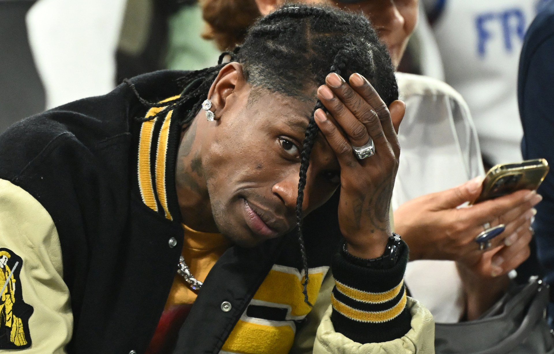 Man with braided hair in a black and gold varsity jacket resting his hand on his head at a basketball game.