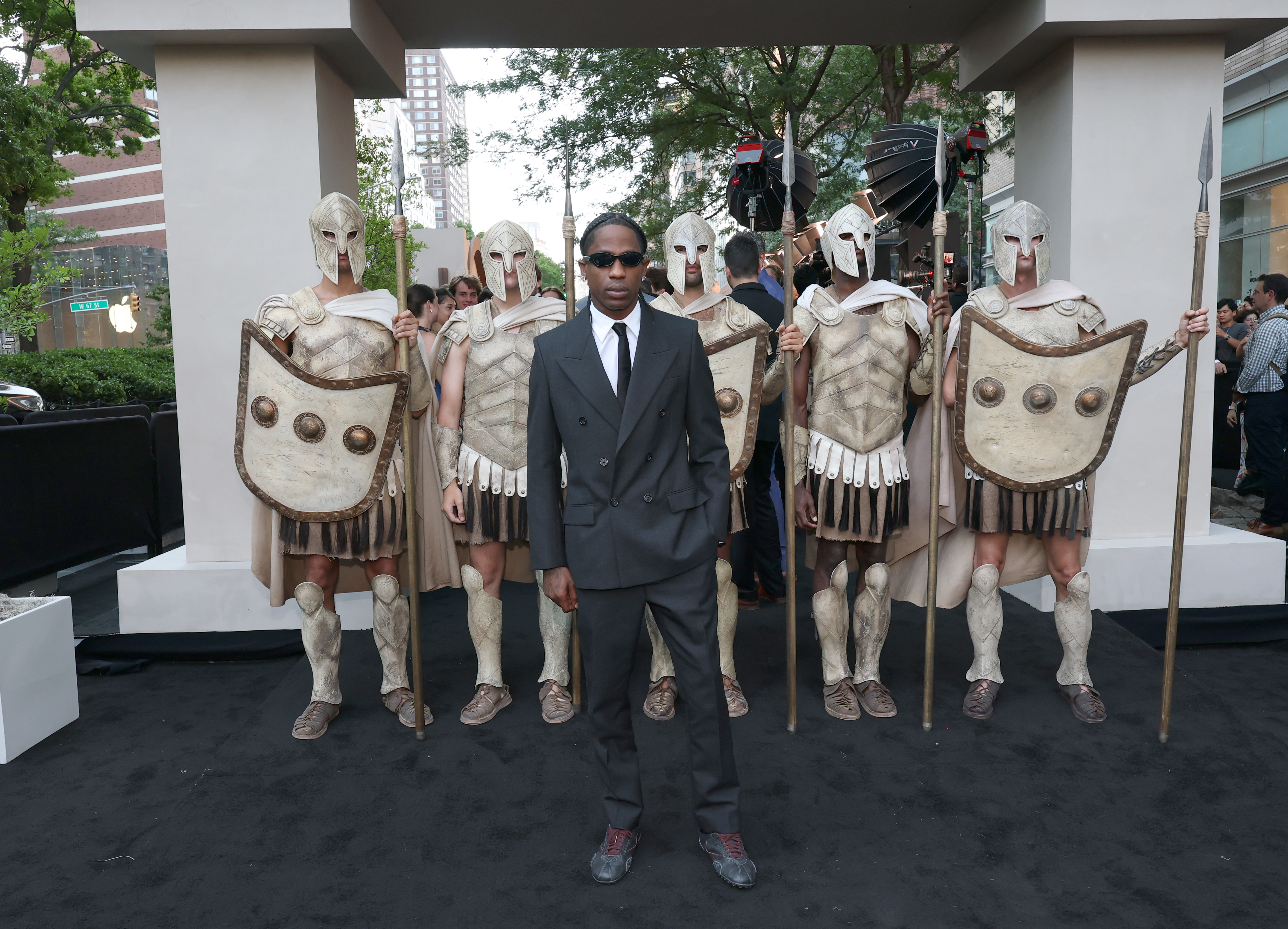 A man in a dark suit and sunglasses standing in front of several performers costumed as Spartan warriors with helmets, shields, and spears at a movie premiere.