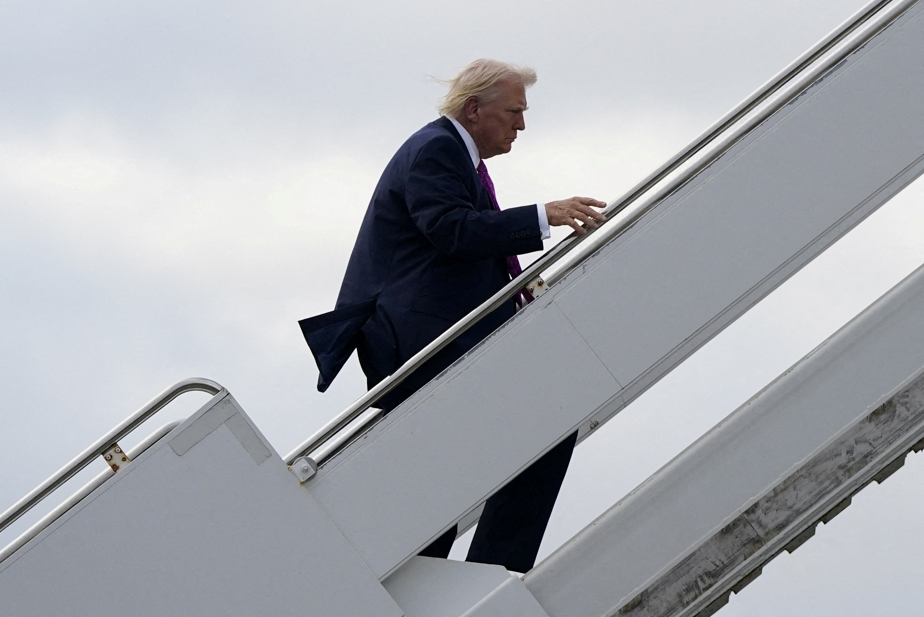 President Donald Trump in a navy suit and purple tie climbing the boarding stairs of Air Force One at an airport, wind blowing his hair.