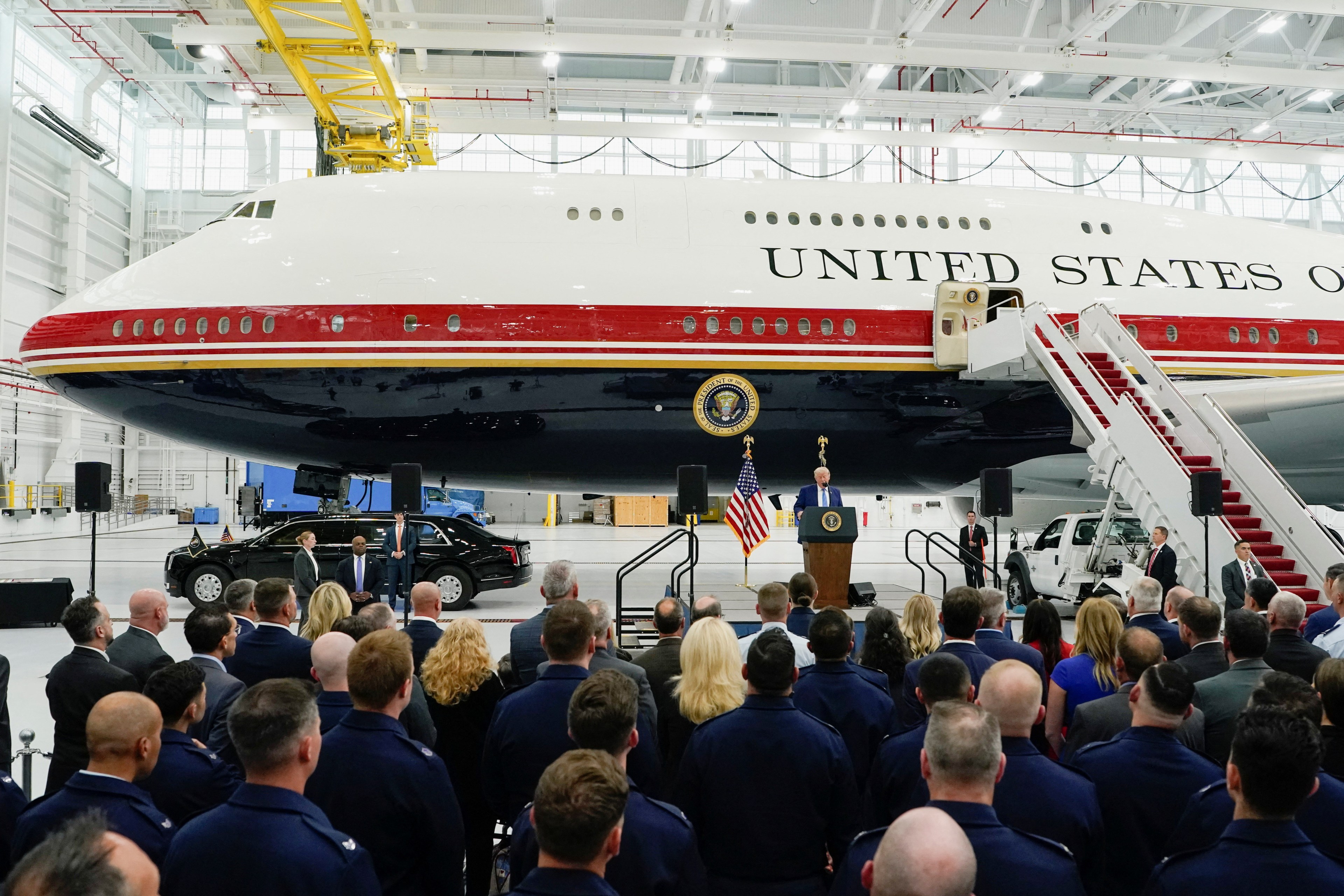 President Trump speaks at a podium before a large crowd inside an aircraft hangar, with a red-and-white Boeing 747 marked 'United States' behind him.