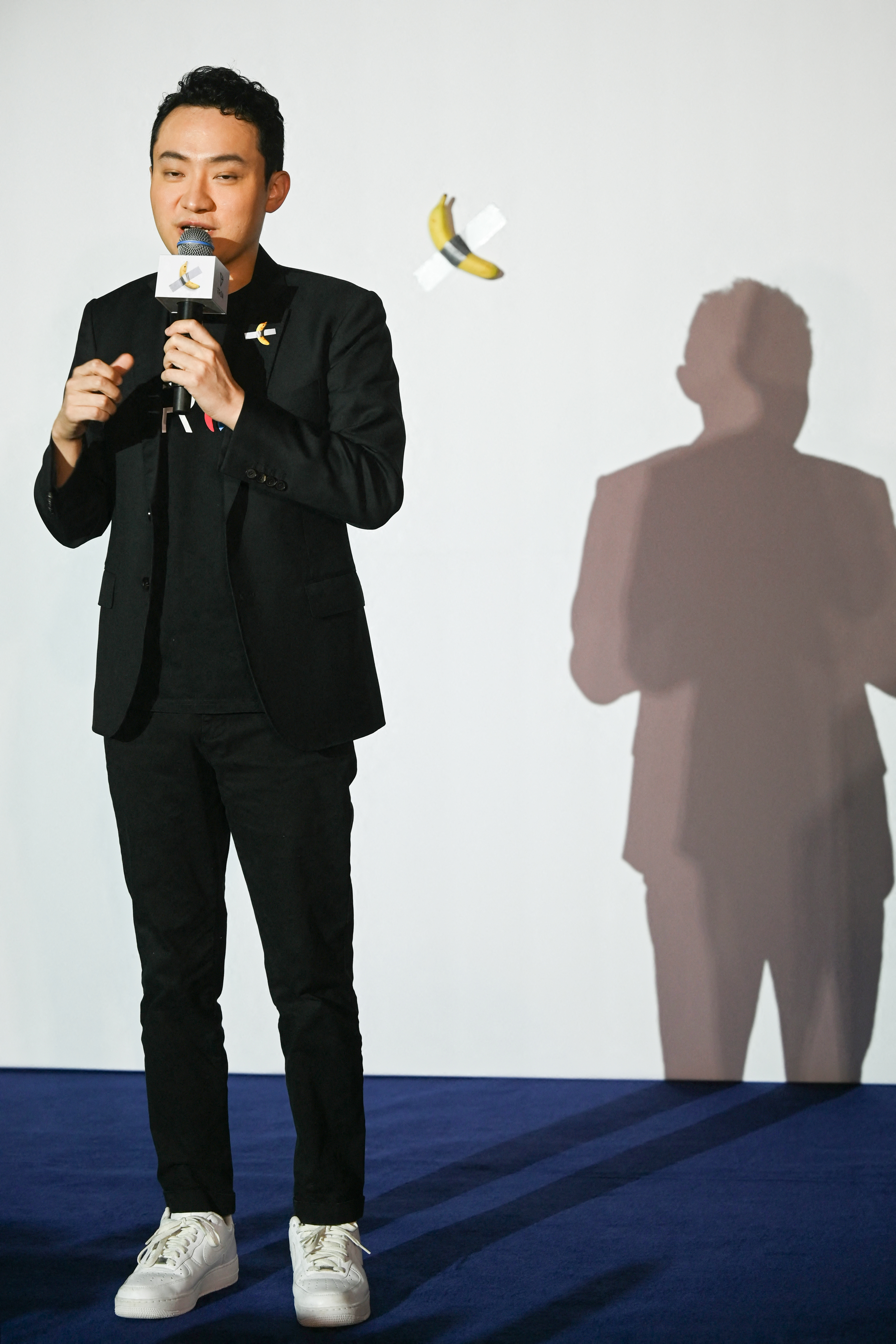 A man in a black suit holds a microphone on stage; behind him, a banana taped to a white wall with duct tape is visible.