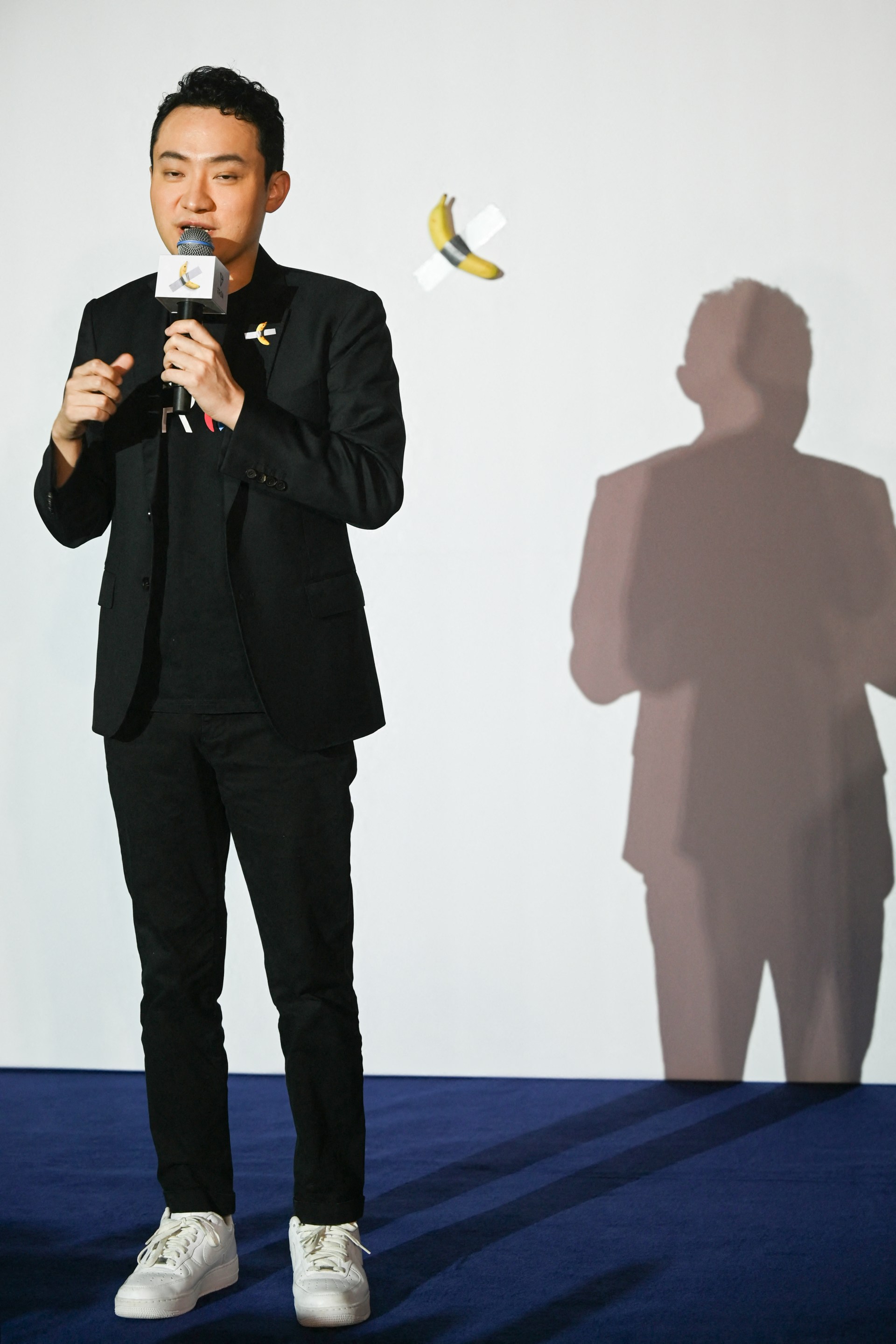 A man in a black suit holds a microphone on stage; behind him, a banana taped to a white wall with duct tape is visible.