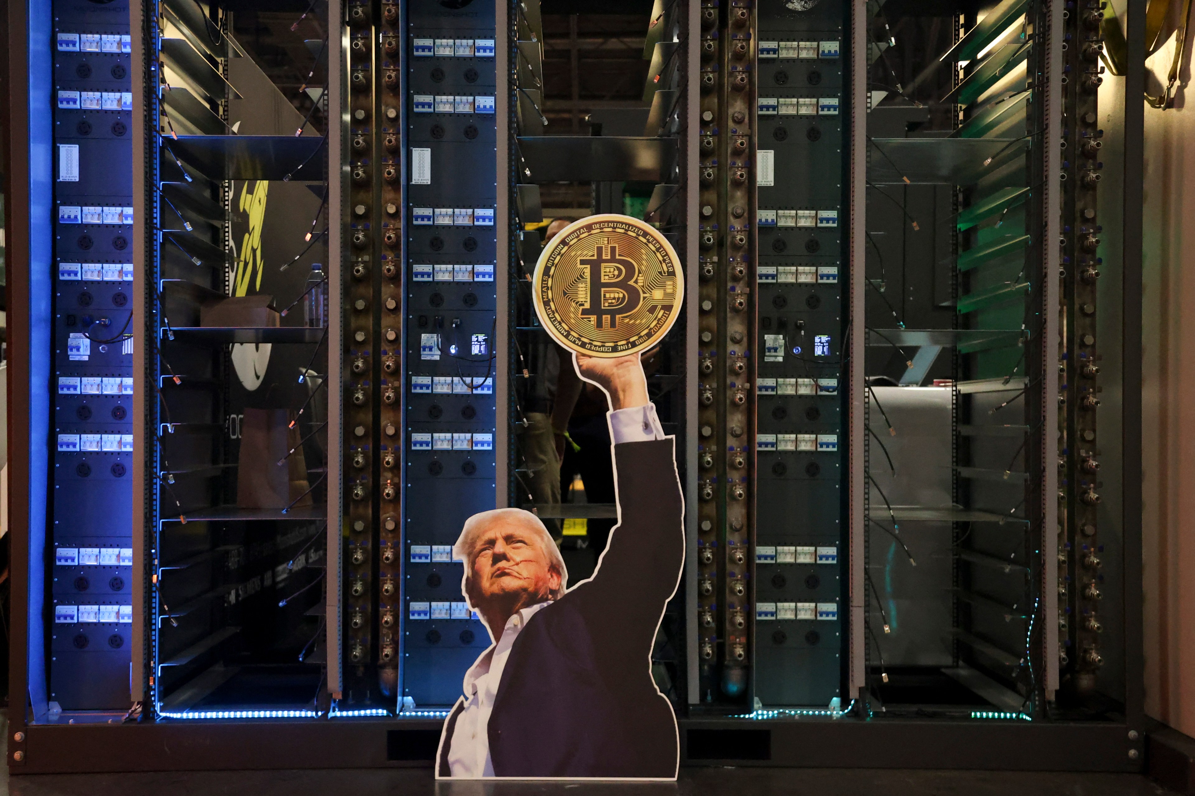 Cardboard cutout of Donald Trump raising a Bitcoin coin, displayed in front of server racks at a crypto conference.