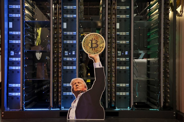 Cardboard cutout of Donald Trump raising a Bitcoin coin, displayed in front of server racks at a crypto conference.