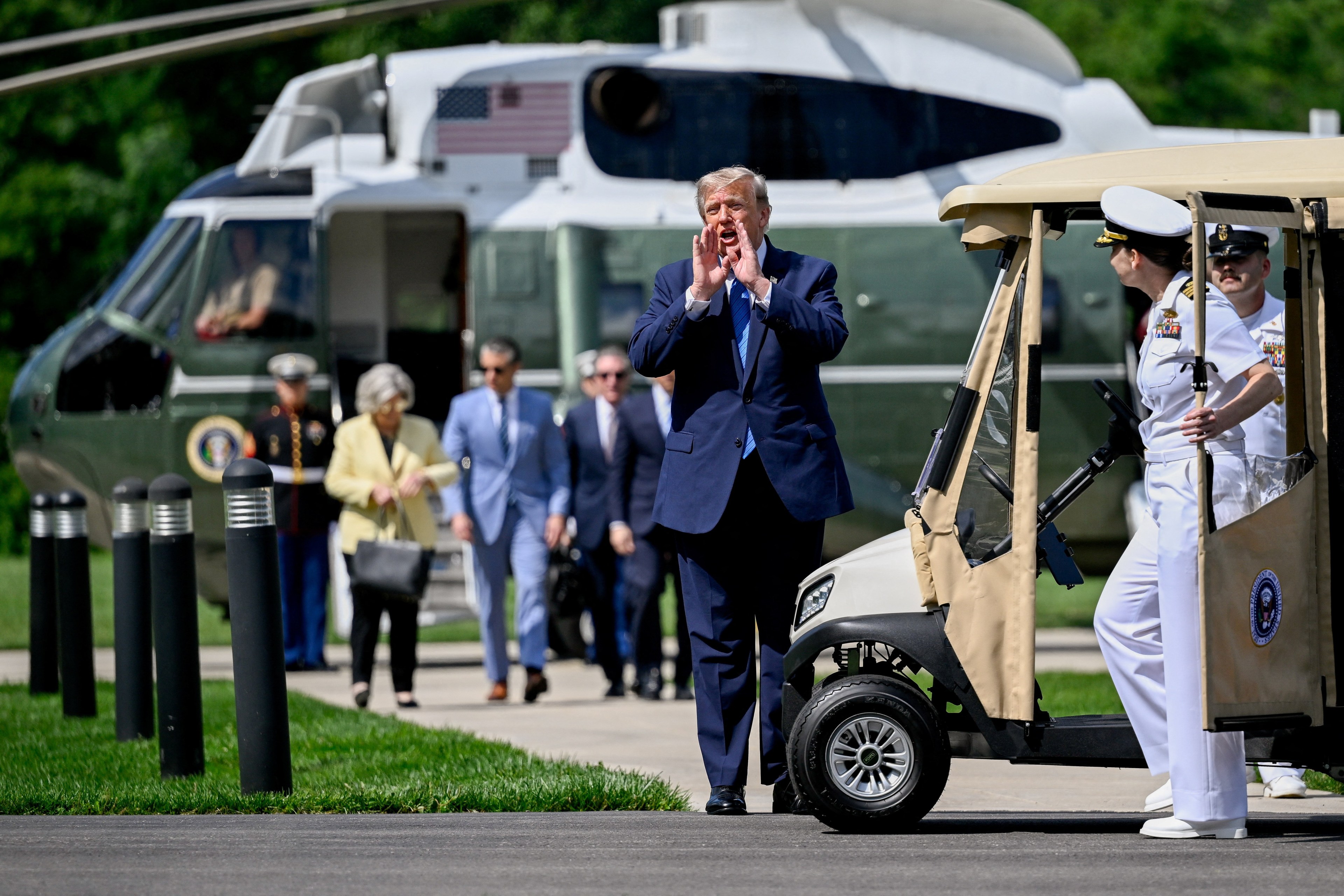 President Trump walks away from Marine One at Camp David, hands cupped around his mouth, with naval aides and a presidential golf cart nearby.