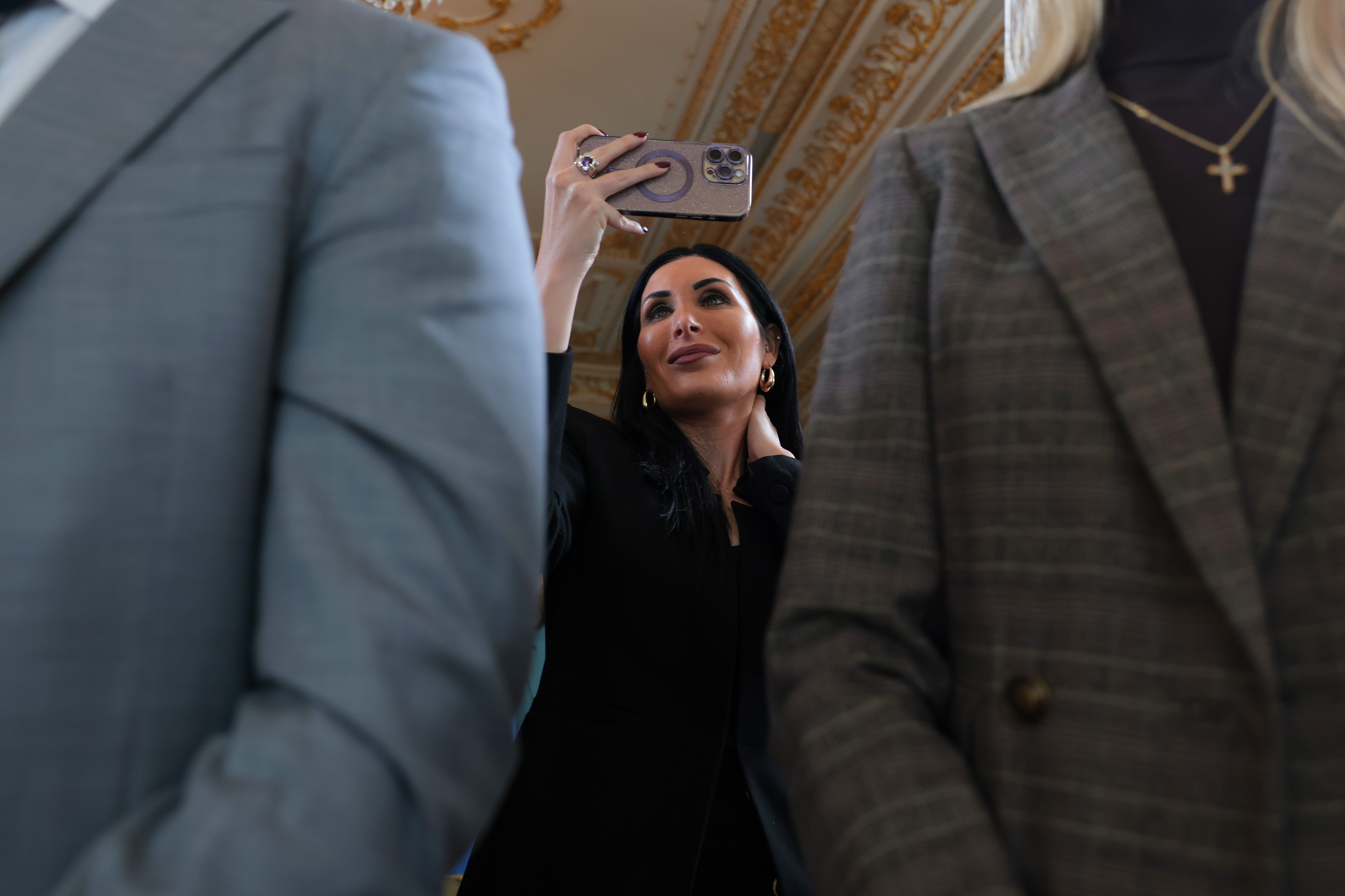 Woman with dark hair in a black outfit holds a smartphone above her head to take a photo inside an ornate gilded room.