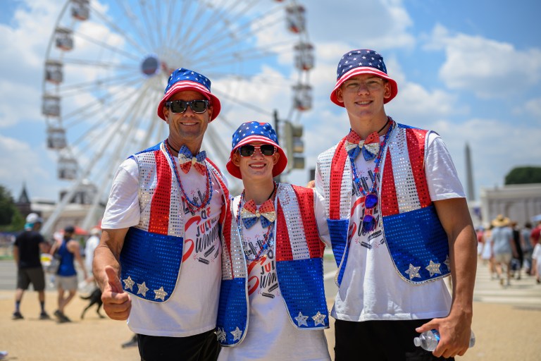 Three people in matching red, white, and blue sequined vests and American flag bucket hats smile in front of a large Ferris wheel on a sunny day.
