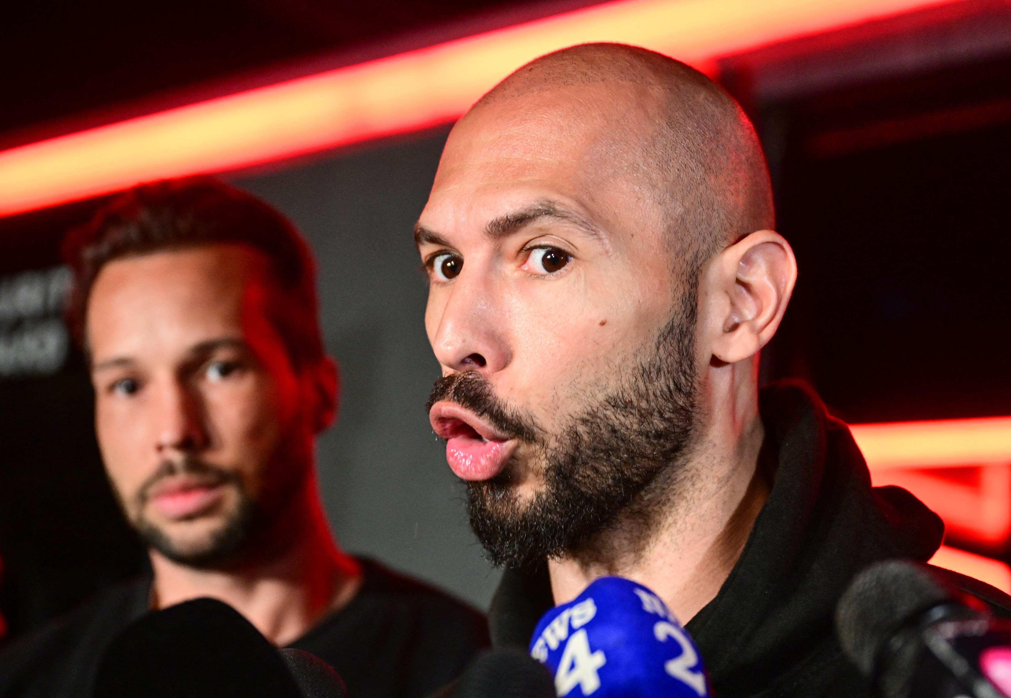 Bald bearded man speaking at a press conference with microphones, another bearded man visible in the background.