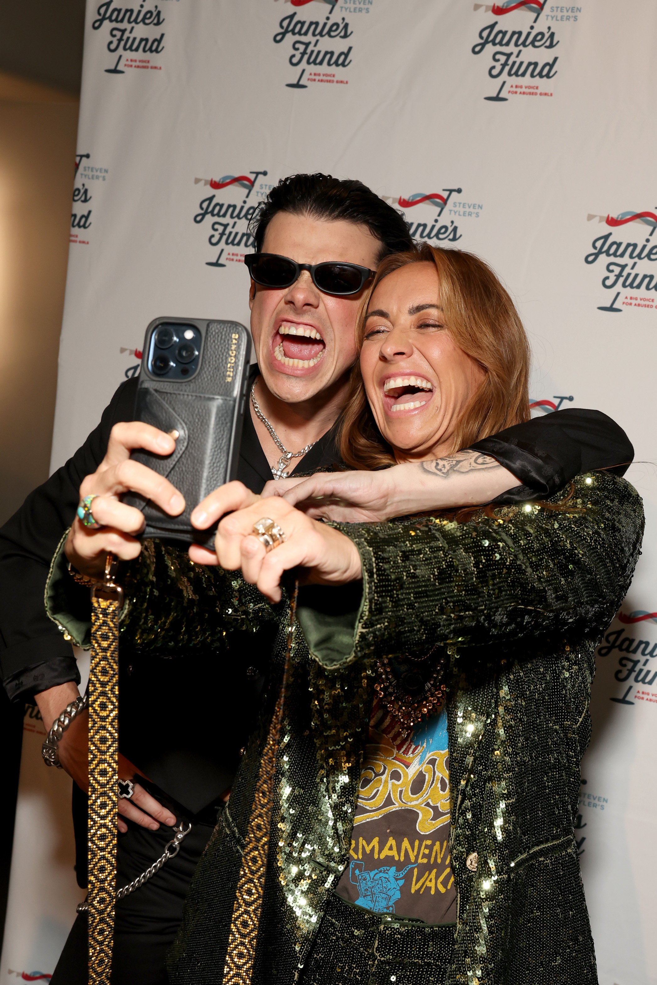 YUNGBLUD in black outfit and sunglasses takes a selfie with Riawna Capri in a green sequined jacket at a Janie's Fund event backdrop.