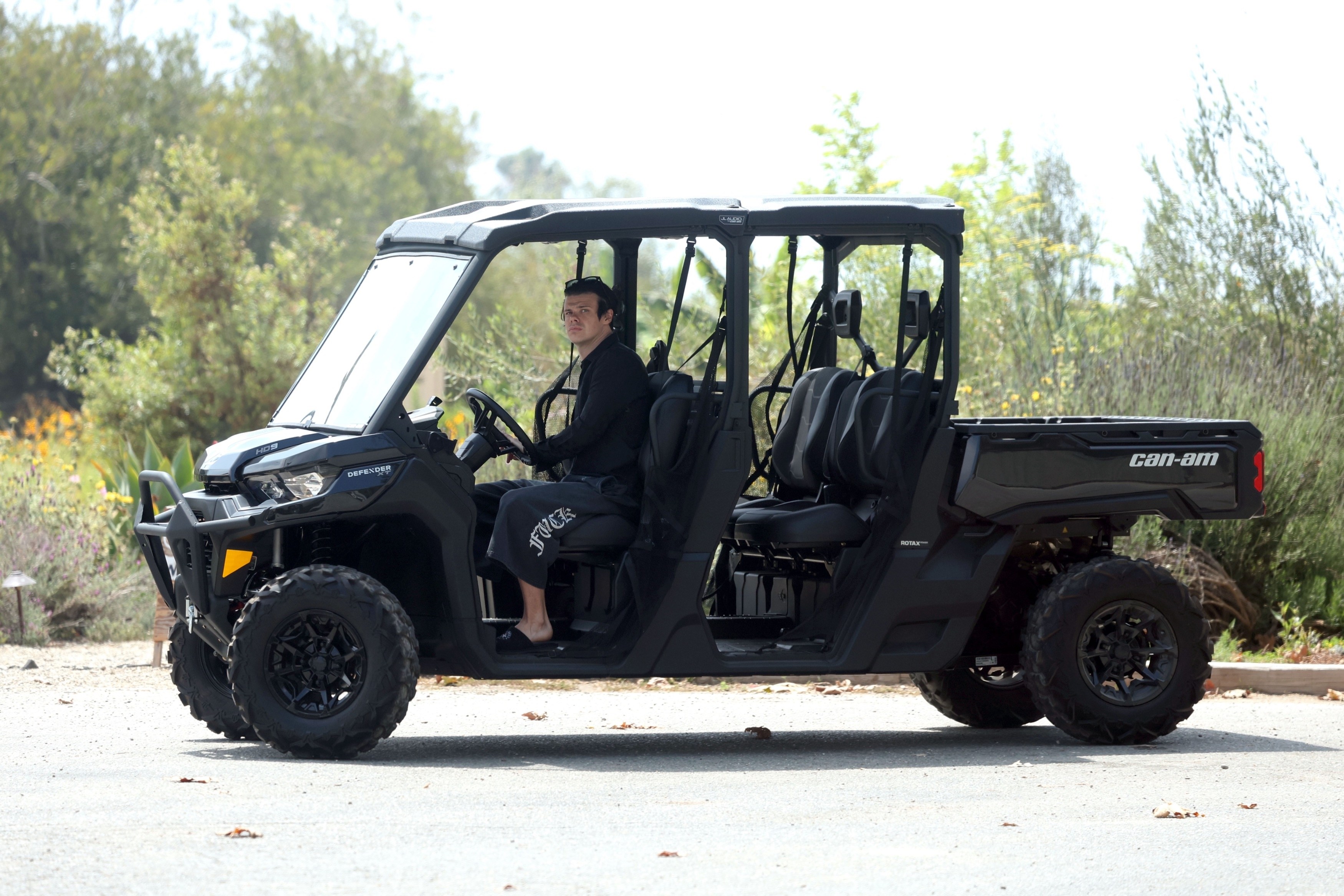 A man in black Chrome Hearts sweatpants and a cropped tee sits barefoot in the driver's seat of a black Can-Am Defender Max side-by-side UTV on a paved path surrounded by shrubs.