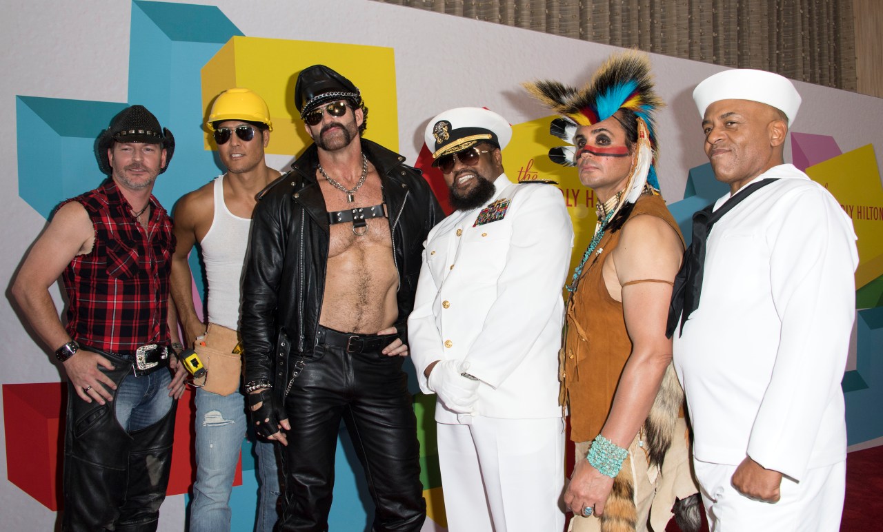 Six Village People members in costume — cowboy, construction worker, biker, navy captain, Native American, and sailor — posing together at a red carpet event.