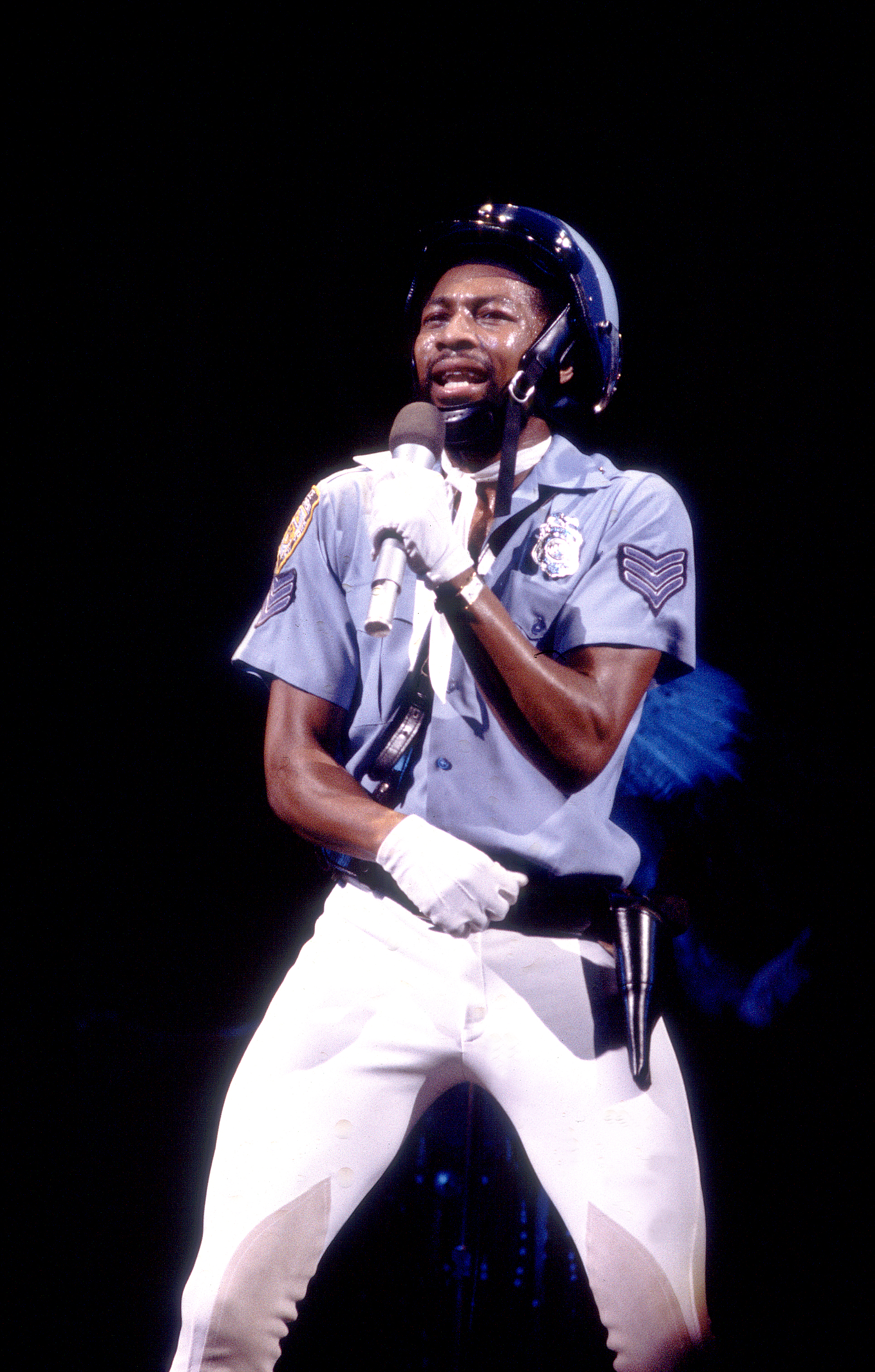 Victor Willis of the Village People performs onstage in a police uniform, helmet, and white gloves, singing into a microphone against a dark background.