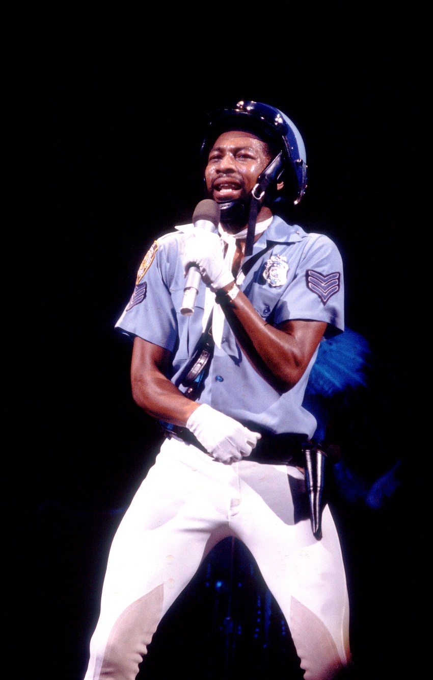 Victor Willis of the Village People performs onstage in a police uniform, helmet, and white gloves, singing into a microphone against a dark background.