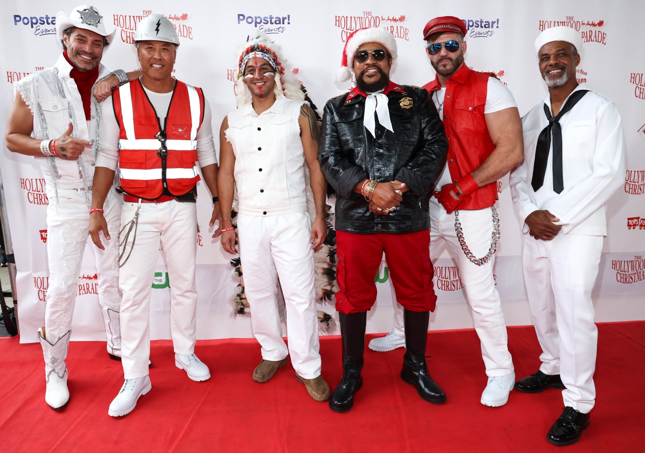 Six men in Village People costumes — cowboy, construction worker, Native American headdress, police-style outfit, biker, and sailor — pose together on a red carpet at the Hollywood Christmas Parade.
